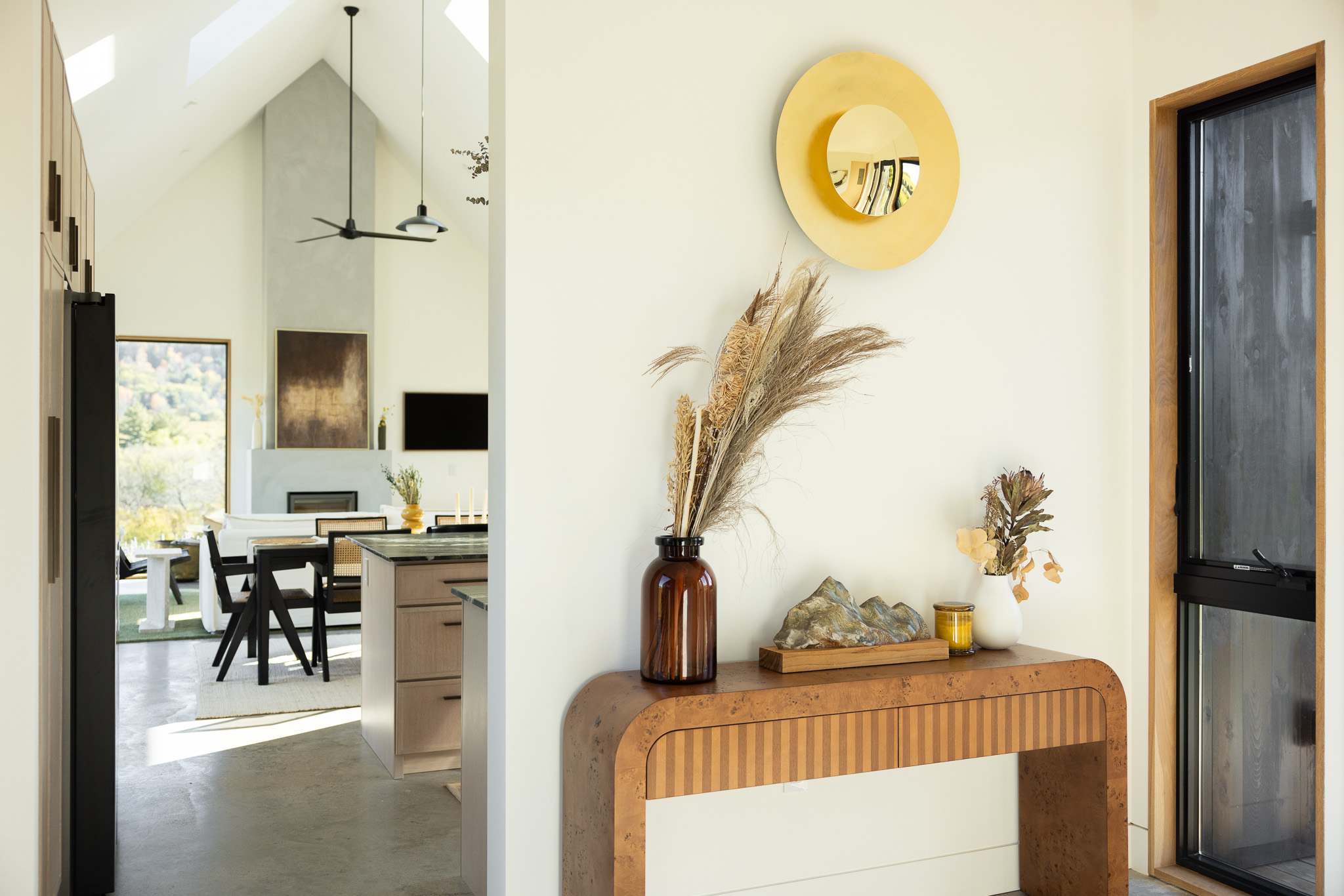 A modern entryway features a curved wooden console table with vertical slat details, topped with a large amber vase holding dried pampas grass and a decorative mountain sculpture. Above, a circular gold mirror reflects the bright, open-concept living space with vaulted ceilings and a fireplace.