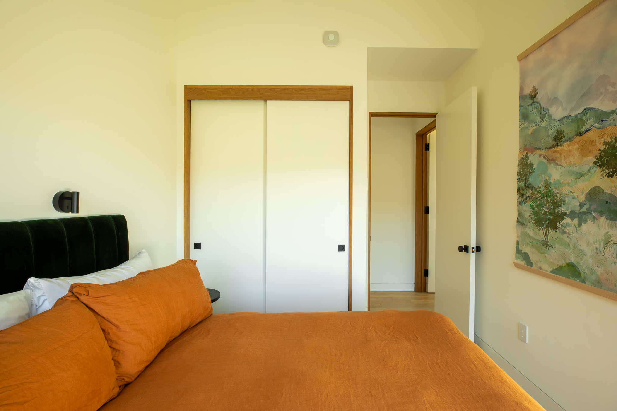View of the bedroom with orange bedding, showing a built-in closet with white sliding doors and a doorway leading to another room, all with light wood trim.