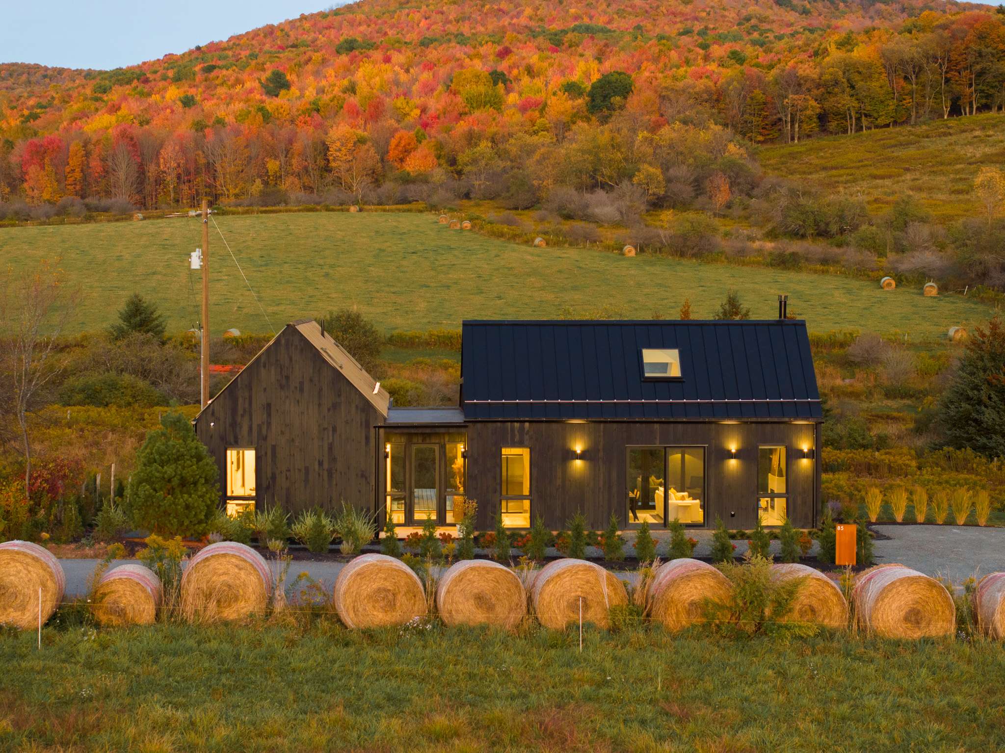Front view of the black modern barn-style house with a dark metal roof, showcasing its clean lines and multiple windows. The house is set against a backdrop of vibrant autumn hills, with hay bales and a gravel driveway in the foreground.