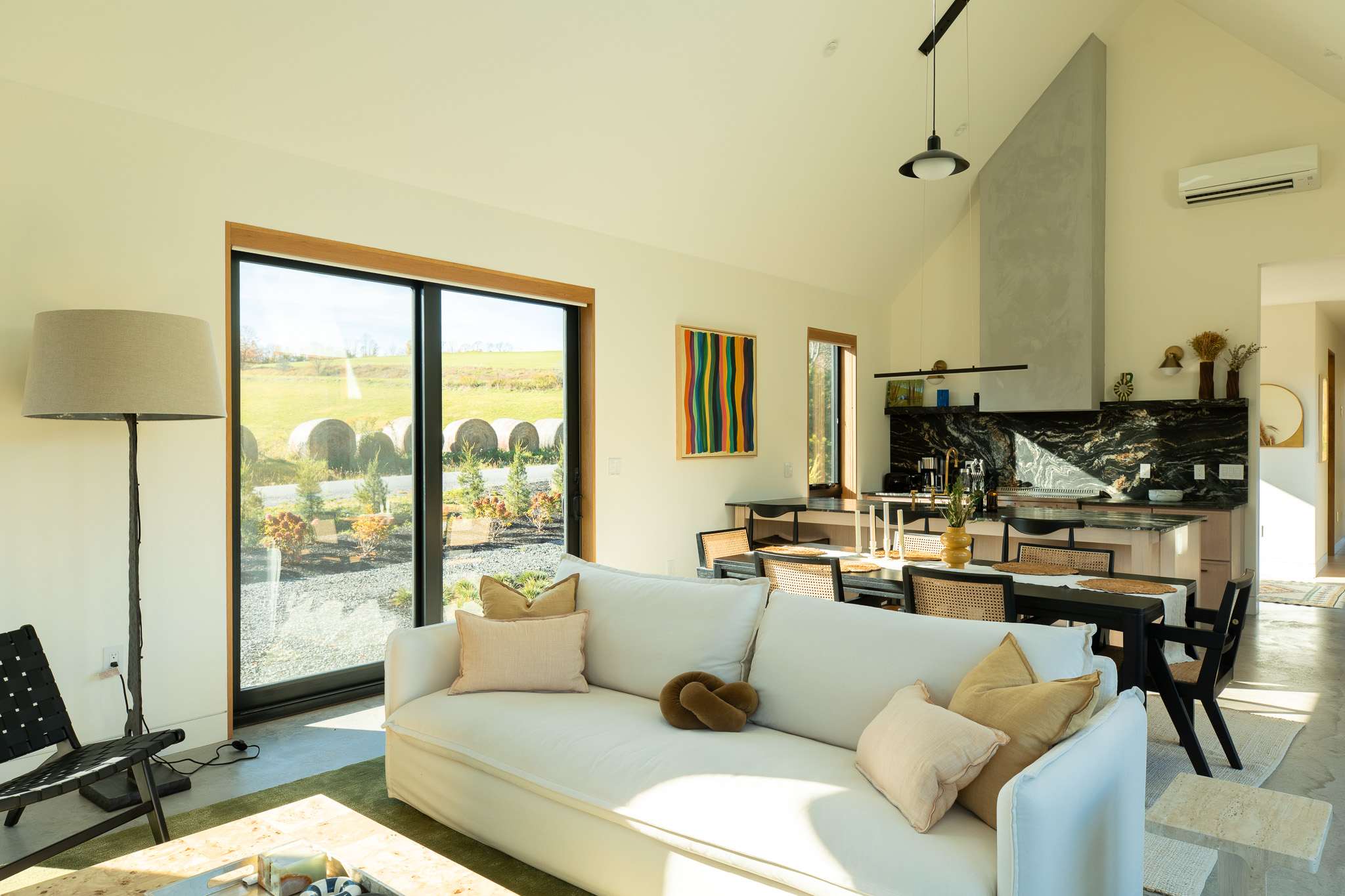 A spacious living room with a white sofa, a floor lamp, and a light-colored coffee table on a green rug. Large sliding glass doors provide abundant natural light and views of the outdoor landscape with hay bales and distant hills.
