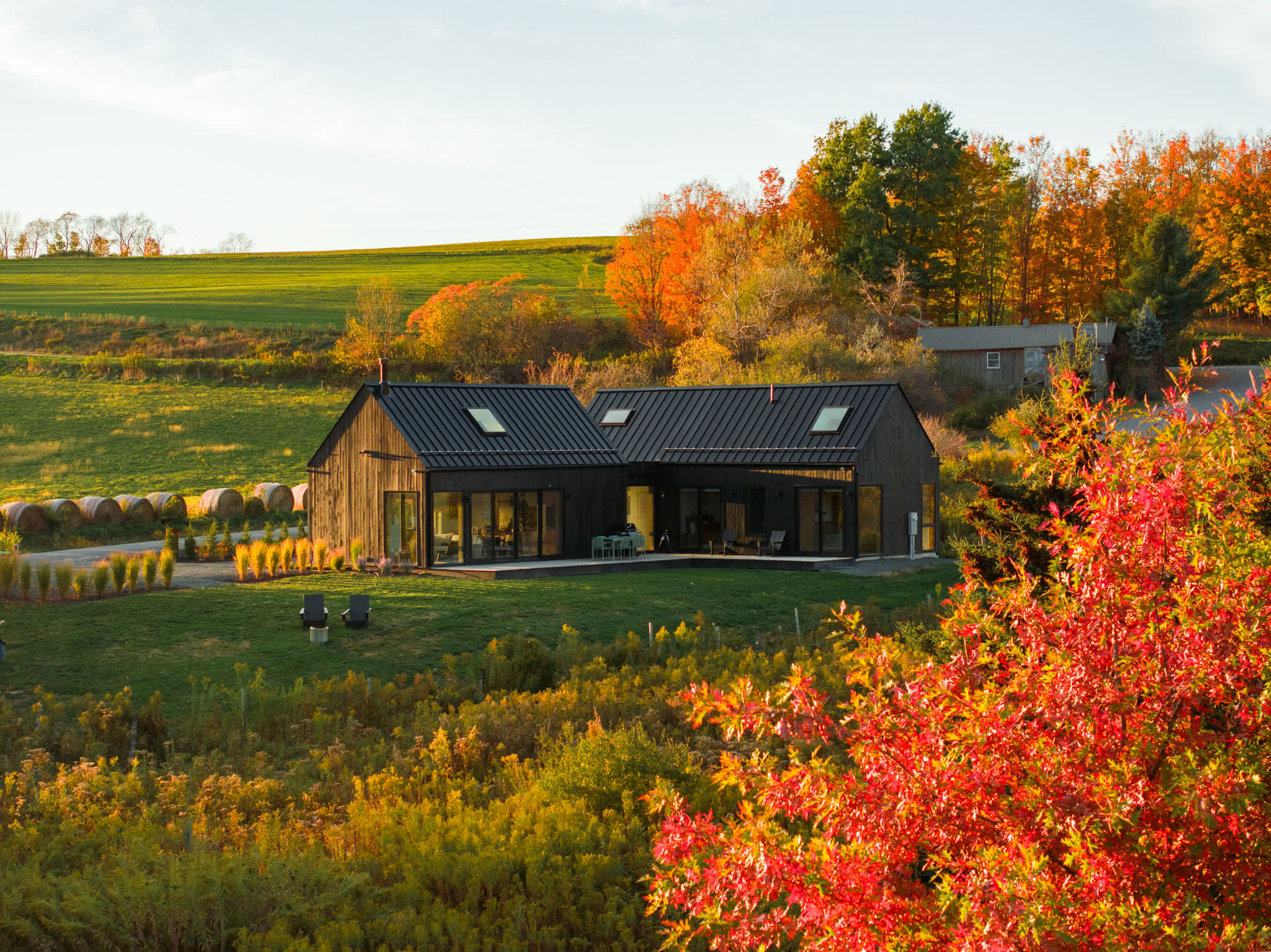 Aerial view of the black modern barn-style house nestled in a vibrant autumn landscape with rolling green and orange hills. The property features a large yard and is surrounded by colorful trees and fields.