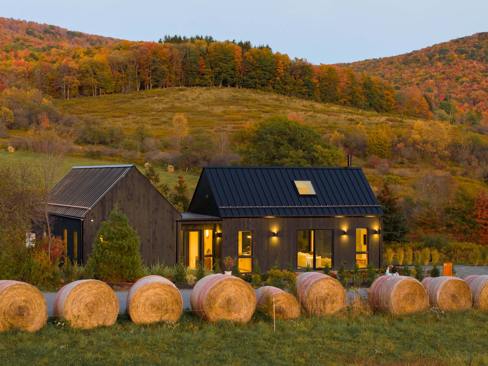 Elevated view of the black modern barn-style house with a dark metal roof, surrounded by a picturesque autumn landscape. Hay bales are neatly arranged in the foreground, leading to the house with a backdrop of vibrant fall-colored hills.