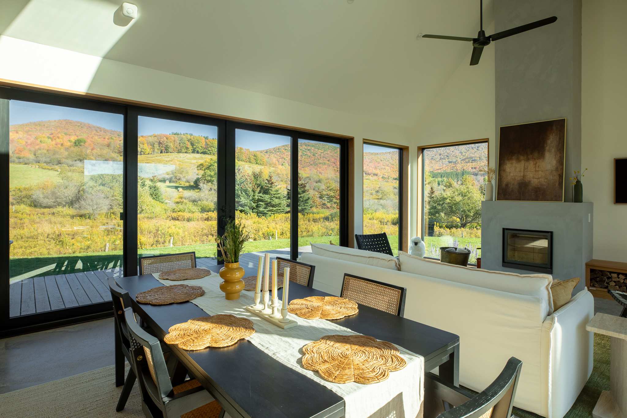 A bright dining area with a black table and cane-backed chairs, set against a backdrop of floor-to-ceiling sliding glass doors that open to a deck and offer expansive views of autumn-colored hills. A modern fireplace and a white sofa are visible in the adjacent living space.