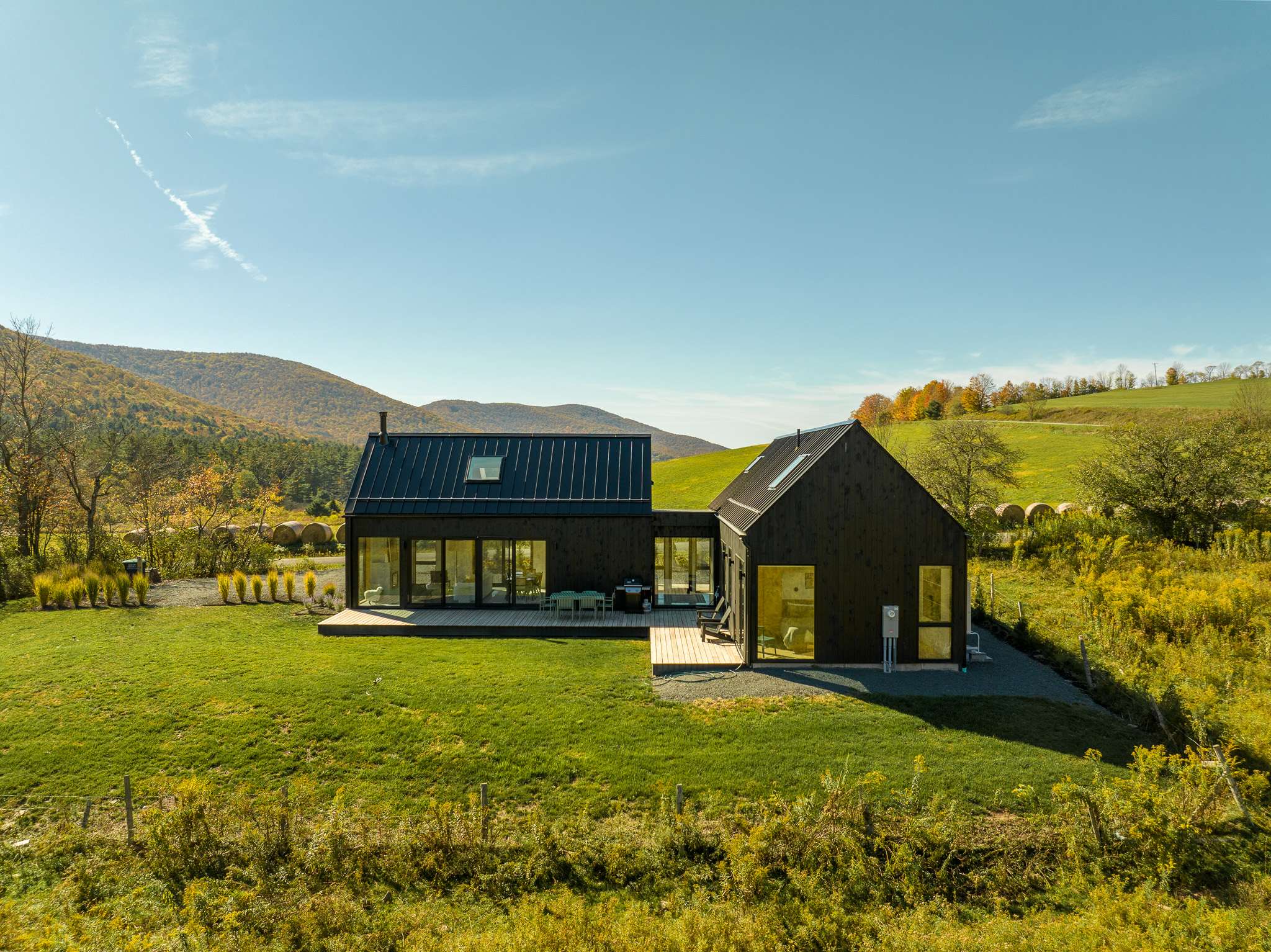 Aerial view of a modern black house with a dark metal roof, featuring multiple large windows and decks, situated in a vast green meadow surrounded by autumn-colored mountains under a clear blue sky.