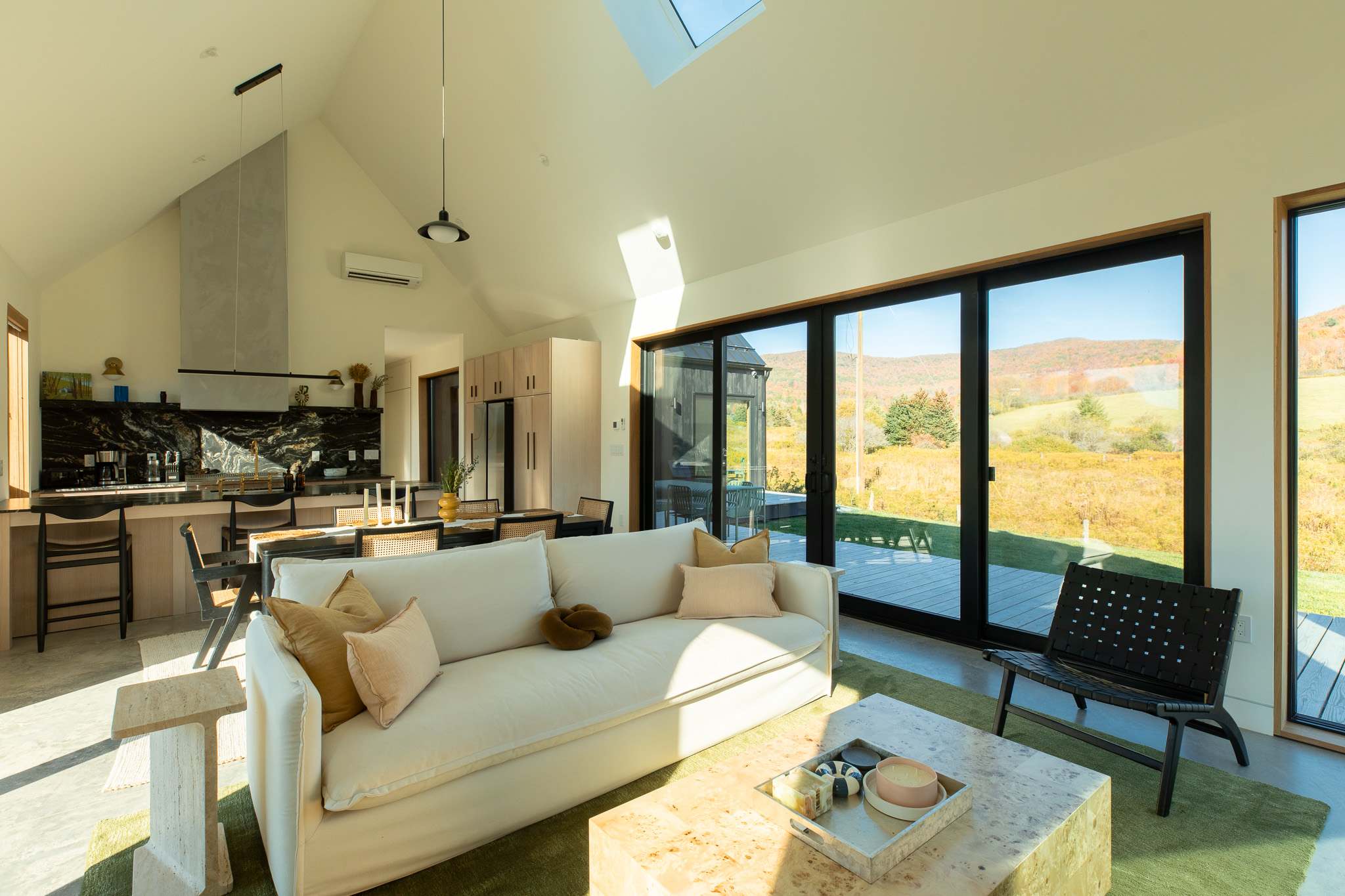 A bright, open-concept living room features a large white sofa with throw pillows, a light-colored coffee table, and a green rug. Floor-to-ceiling sliding glass doors offer panoramic views of the rolling hills, connecting the indoor space with the natural surroundings.