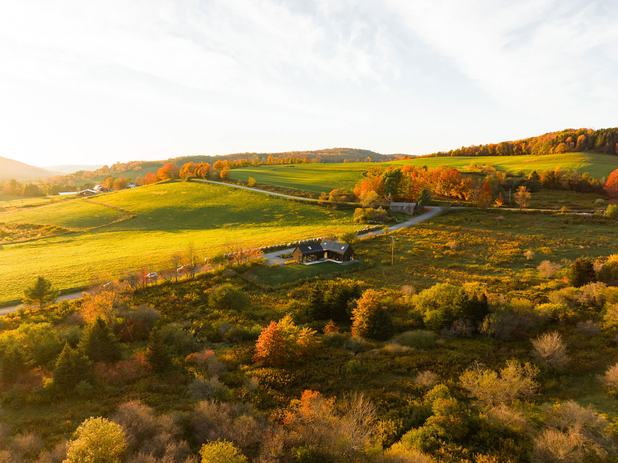 Expansive aerial view showcasing the modern black house nestled within a vast, vibrant autumn landscape of rolling green and golden hills, surrounded by colorful trees and a winding road.