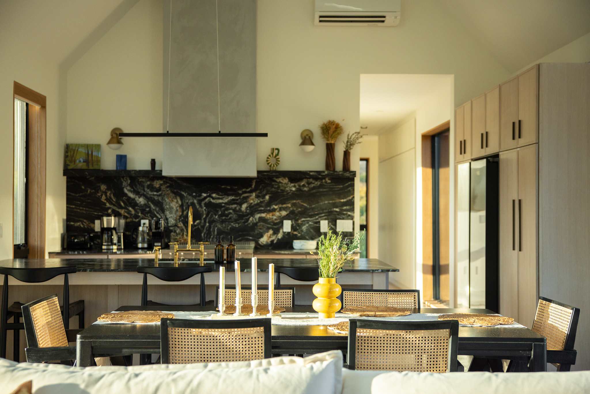 The dining area, viewed from the living room, features a black dining table with cane-backed chairs, set with placemats and a yellow vase. In the background, the open-concept kitchen boasts dark marble countertops and light wood cabinets, illuminated by natural light.