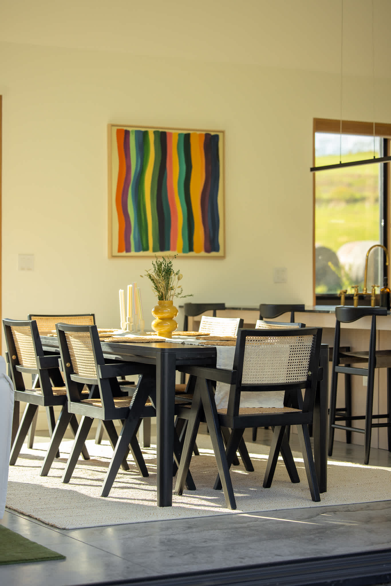 A close-up of the dining area shows a black table with cane-backed chairs, set with a yellow vase and candles. A colorful abstract painting hangs on the wall, and a large window provides a glimpse of the outdoor landscape.