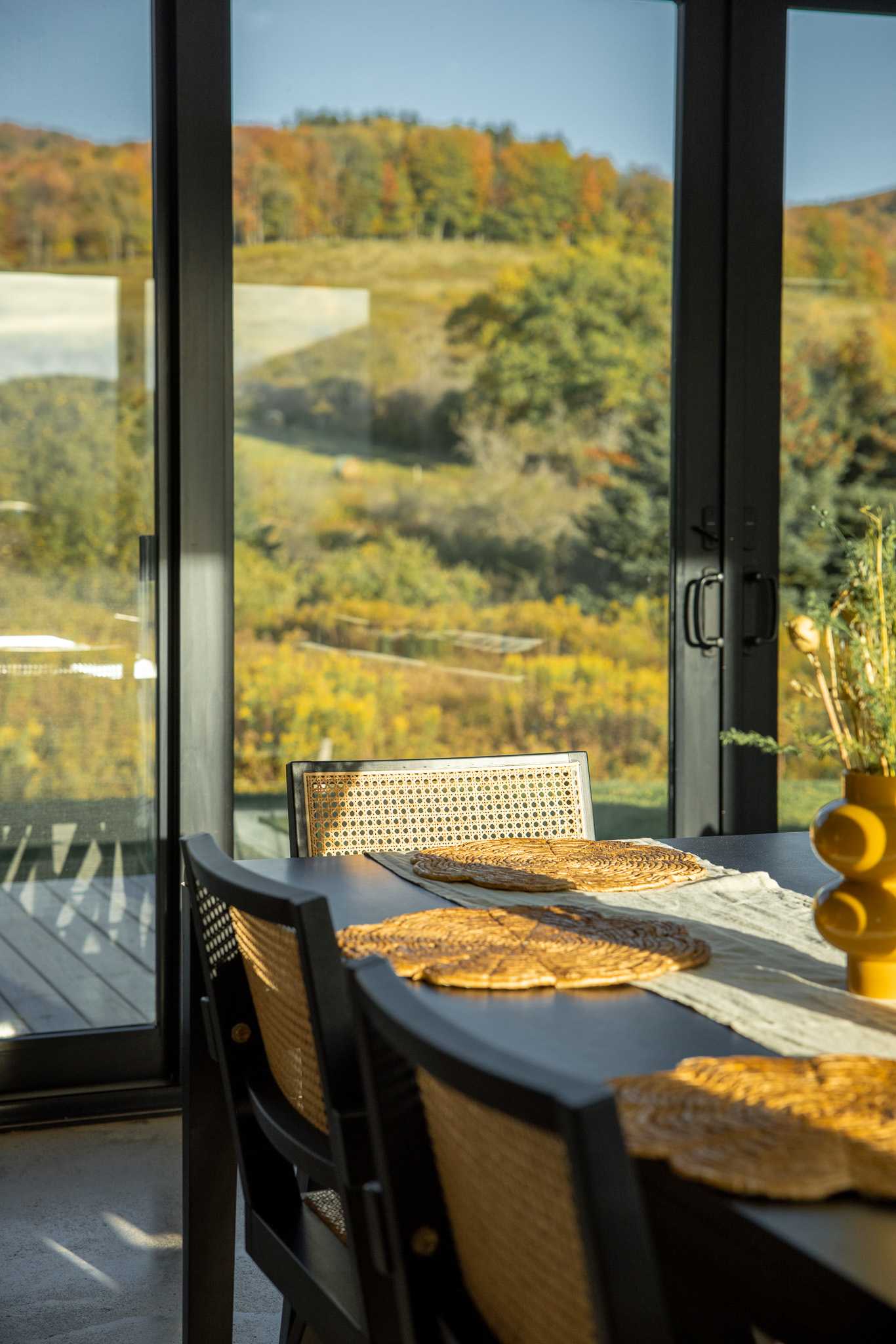 Dining area with a dark wooden table and black chairs featuring cane backs, positioned by large glass doors that offer expansive views of the sun-drenched autumn landscape and distant hills.