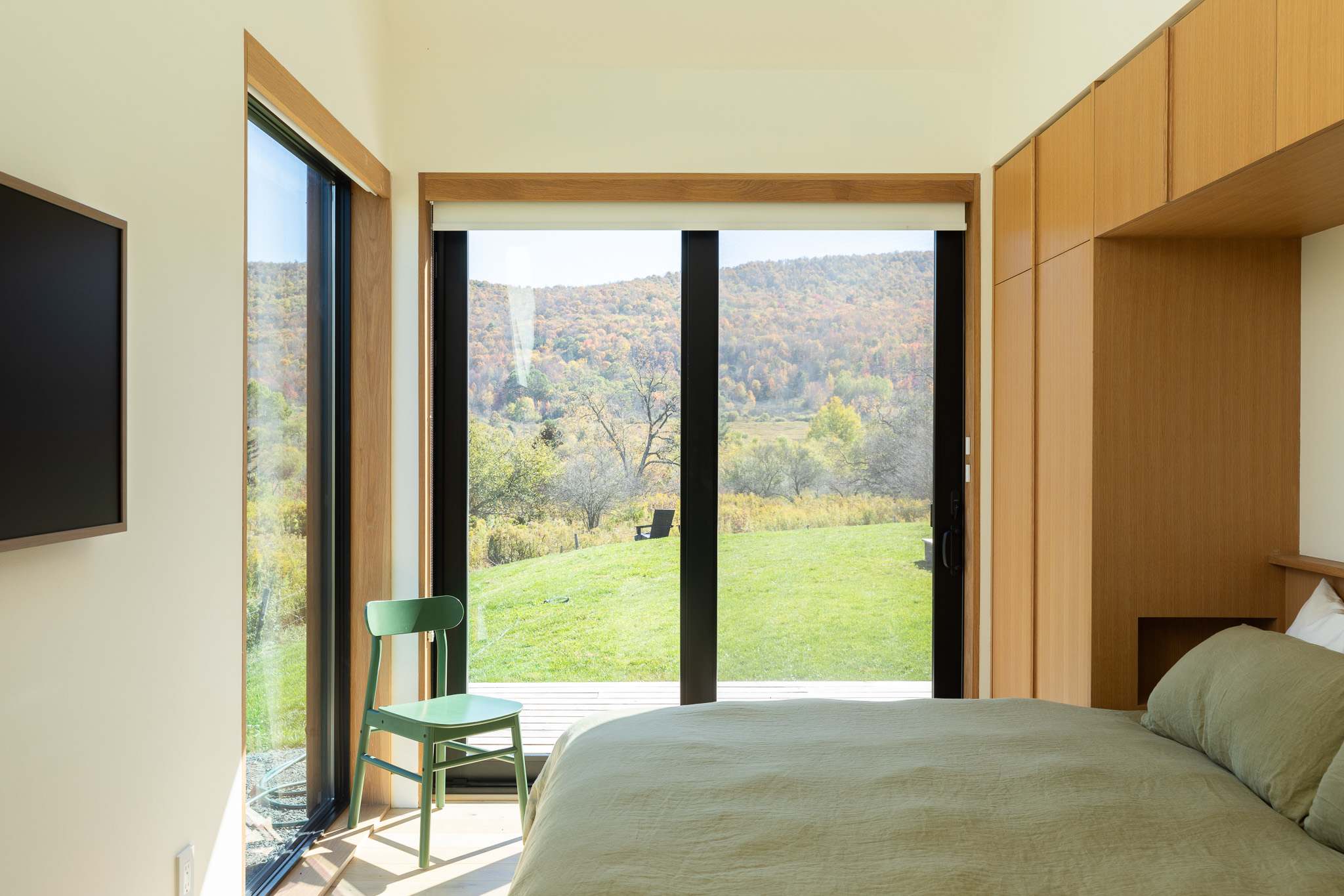 Bedroom with a light green bed, a green wooden chair, and floor-to-ceiling glass doors opening to a deck and offering panoramic views of rolling hills and autumn foliage.