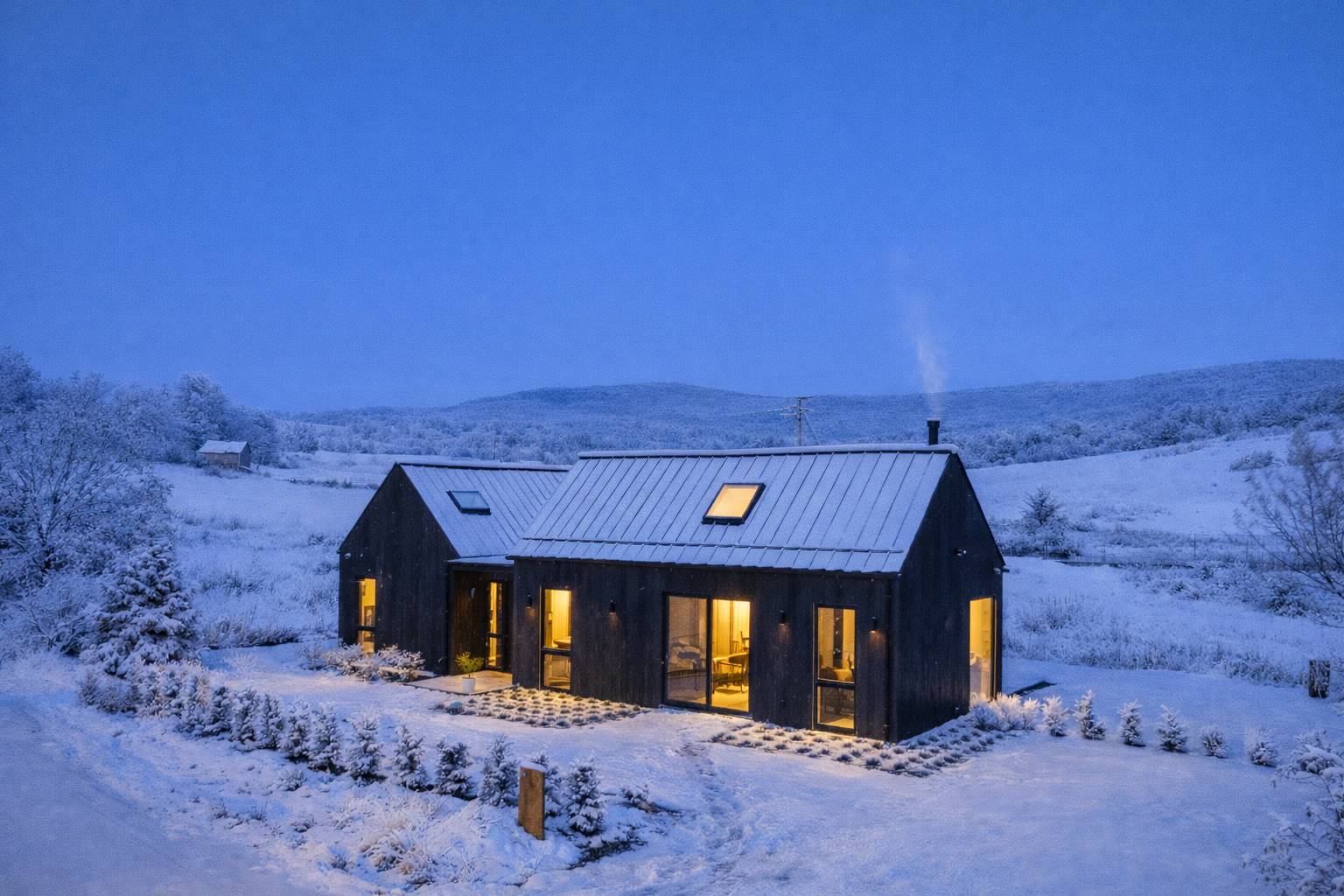 Aerial view of the black modern barn-style house covered in a blanket of snow, with warm lights emanating from its windows against a twilight blue sky. The surrounding landscape is also snow-covered, with distant hills.