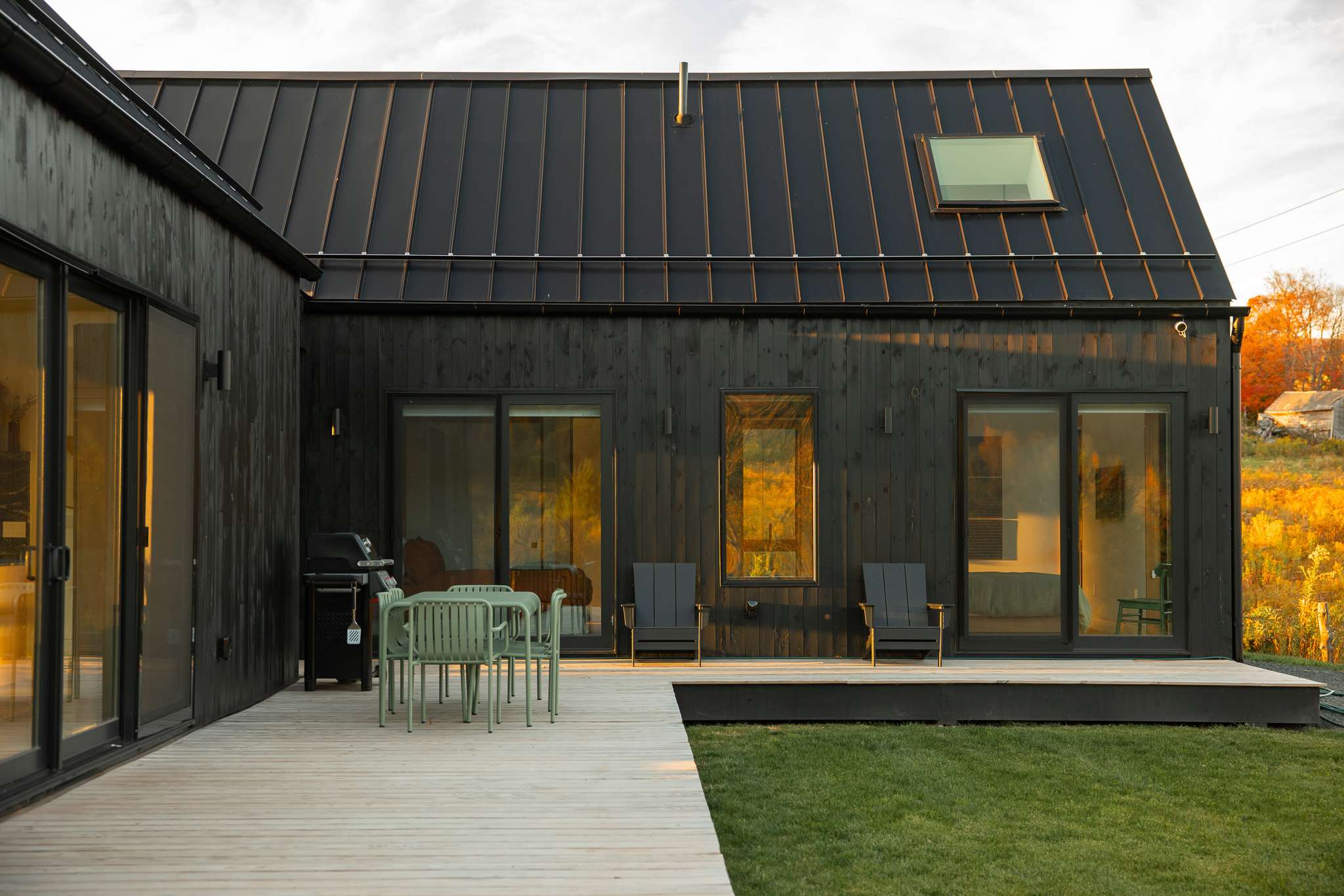 Modern black barn-style house with a dark metal roof and a light wood deck. Features large sliding glass doors, a skylight, a BBQ grill, and an outdoor dining set, overlooking a grassy yard and distant fields.