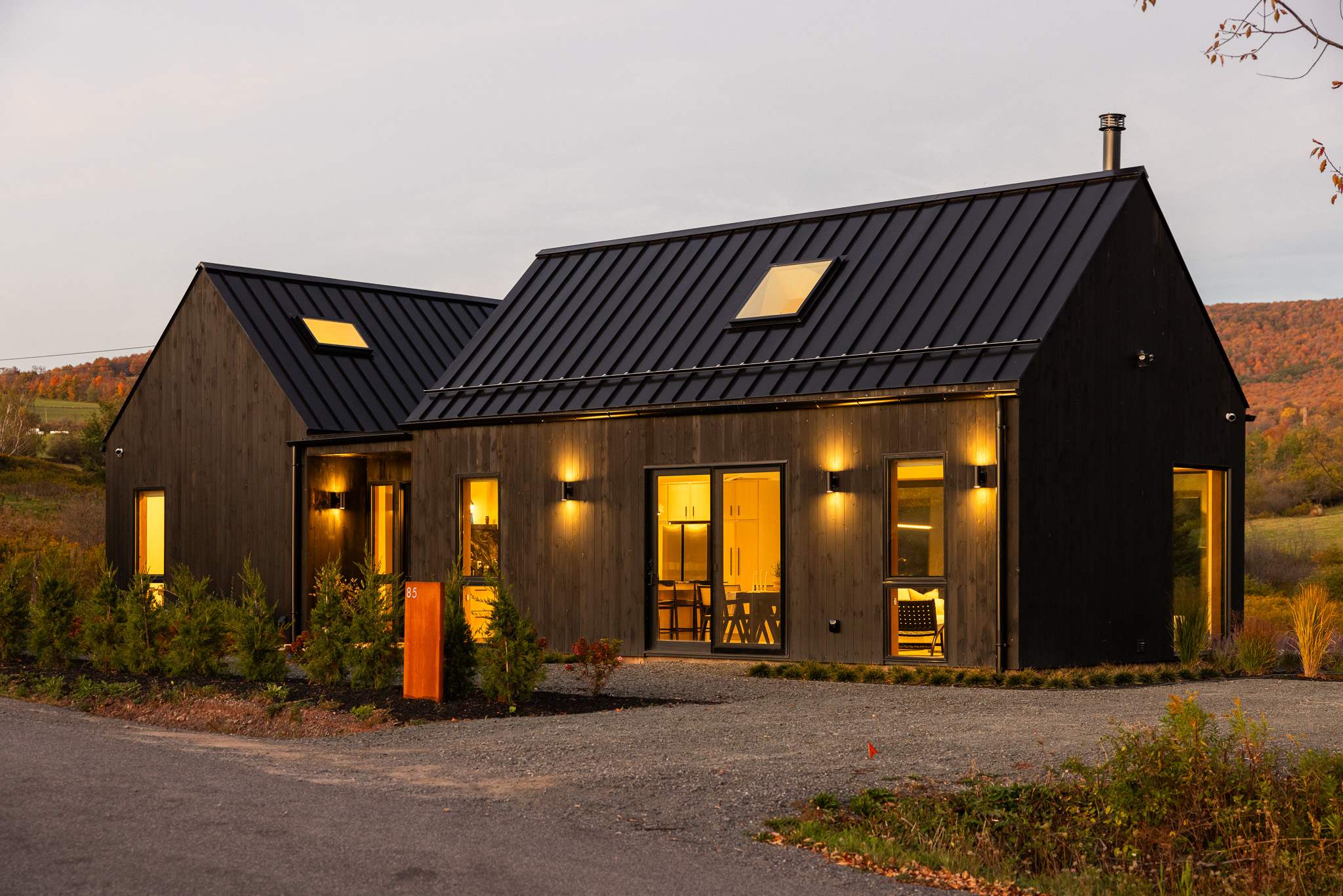 Modern black barn-style house with a dark metal roof and warm interior lights glowing through multiple windows and glass doors at dusk. The exterior features wall-mounted lights and a gravel driveway, with autumn hills in the background.