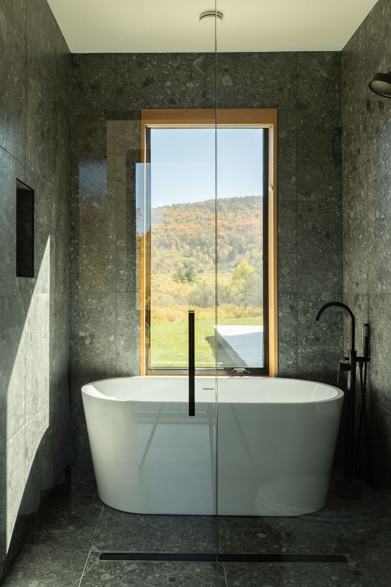 Modern bathroom featuring a freestanding white tub within a glass-enclosed shower, surrounded by dark speckled tile walls and a large window with mountain views.