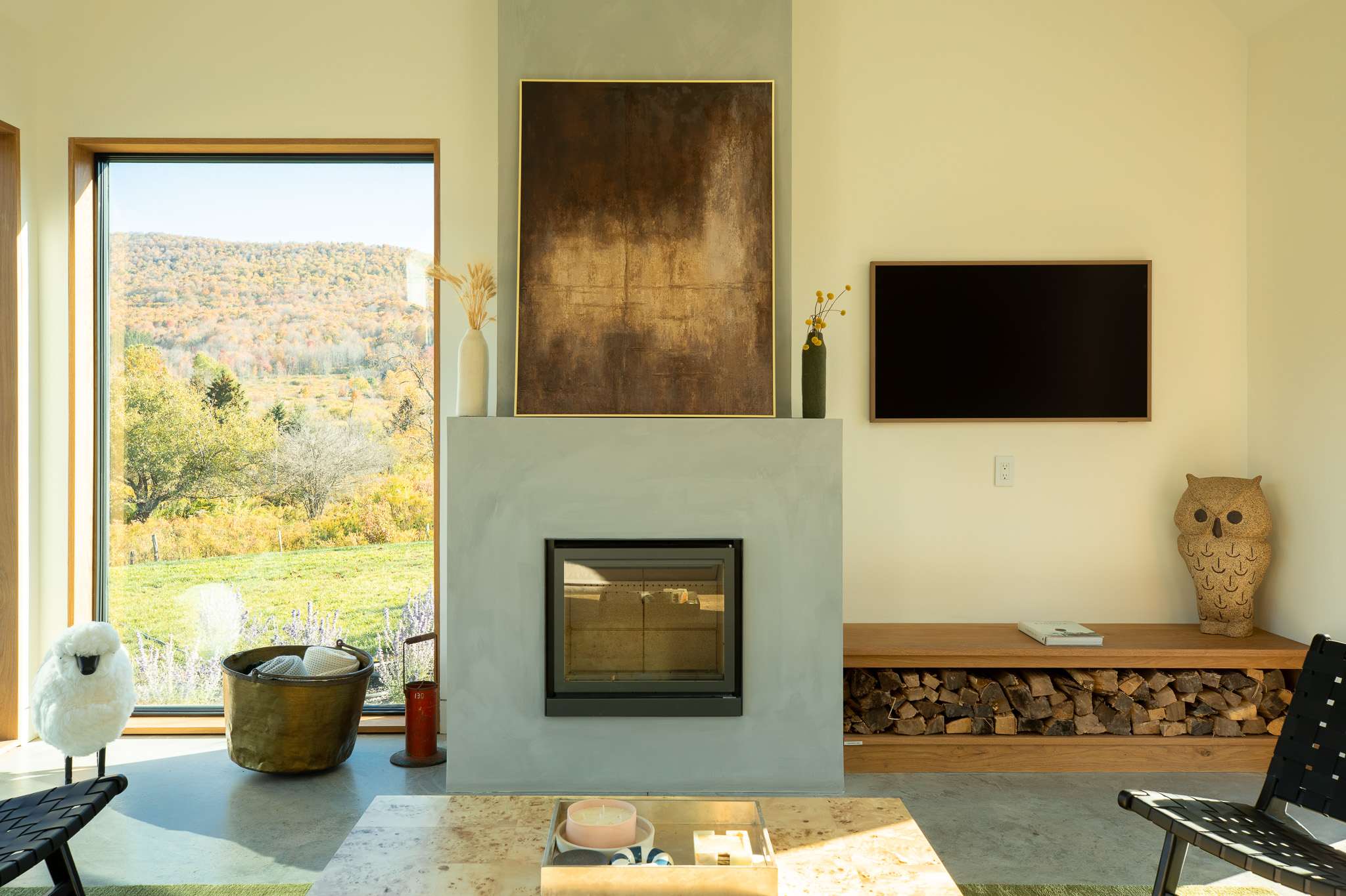 Close-up of the living room's concrete fireplace with a modern insert, flanked by a wooden bench with stacked firewood and a large abstract artwork above.