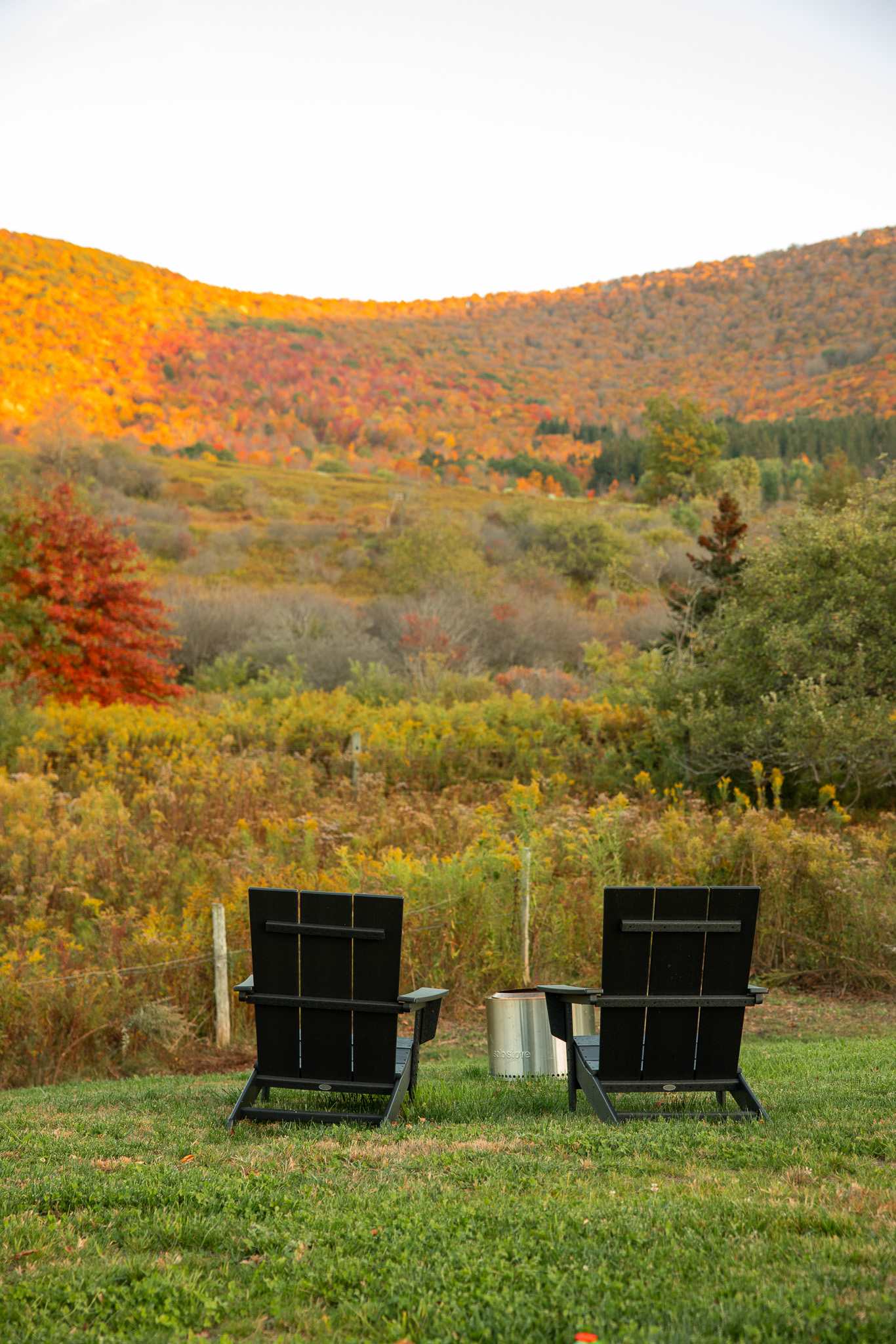 Two black Adirondack chairs and a metal fire pit are set on a grassy lawn, facing a breathtaking view of rolling hills covered in vibrant autumn foliage under a clear sky.
