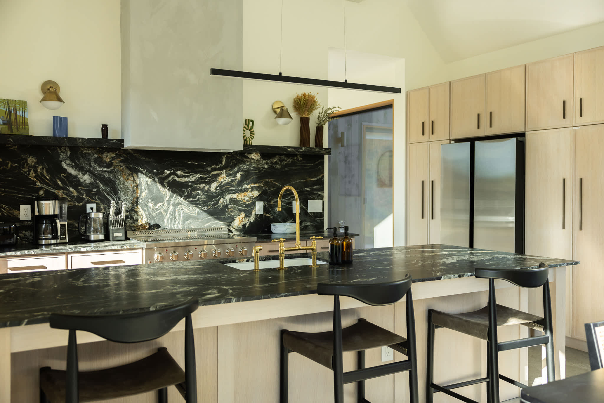 A spacious, modern kitchen with light wood cabinetry and a large island featuring dark marble countertops and three black bar stools. A gold faucet stands out against the dark backsplash, and a stainless steel refrigerator is integrated into the cabinetry.