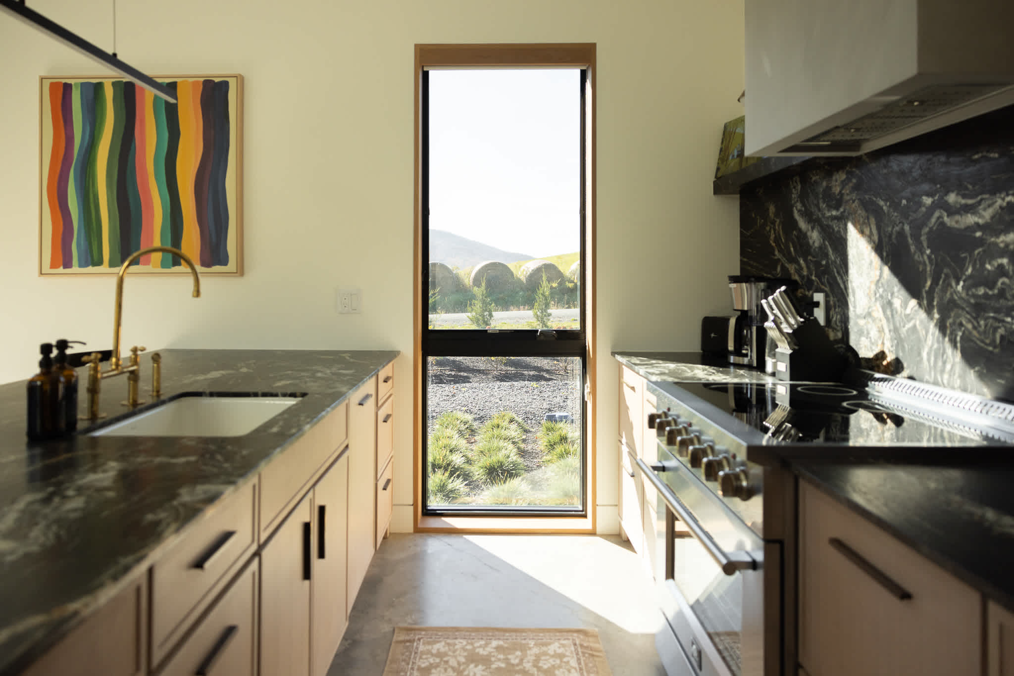 A modern kitchen showcases light wood cabinetry, dark marble countertops, and a matching backsplash with striking white veining. A tall, narrow window offers a view of rolling hills and hay bales, while a gold faucet adds a touch of elegance to the sink area.