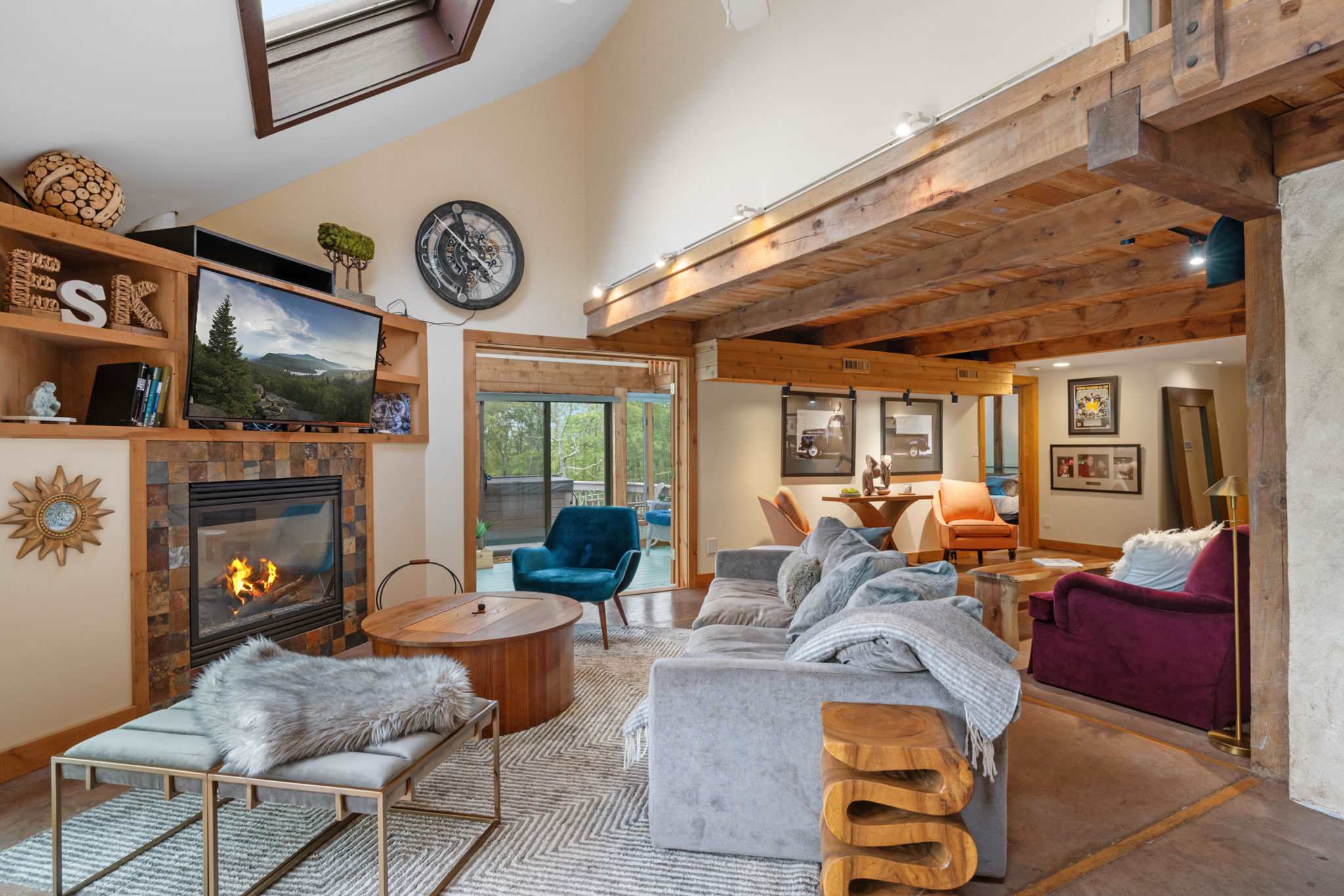 A spacious living room with a vaulted ceiling, exposed wooden beams, and a stone-faced fireplace with a mounted TV, furnished with a large gray sectional sofa and various accent chairs.