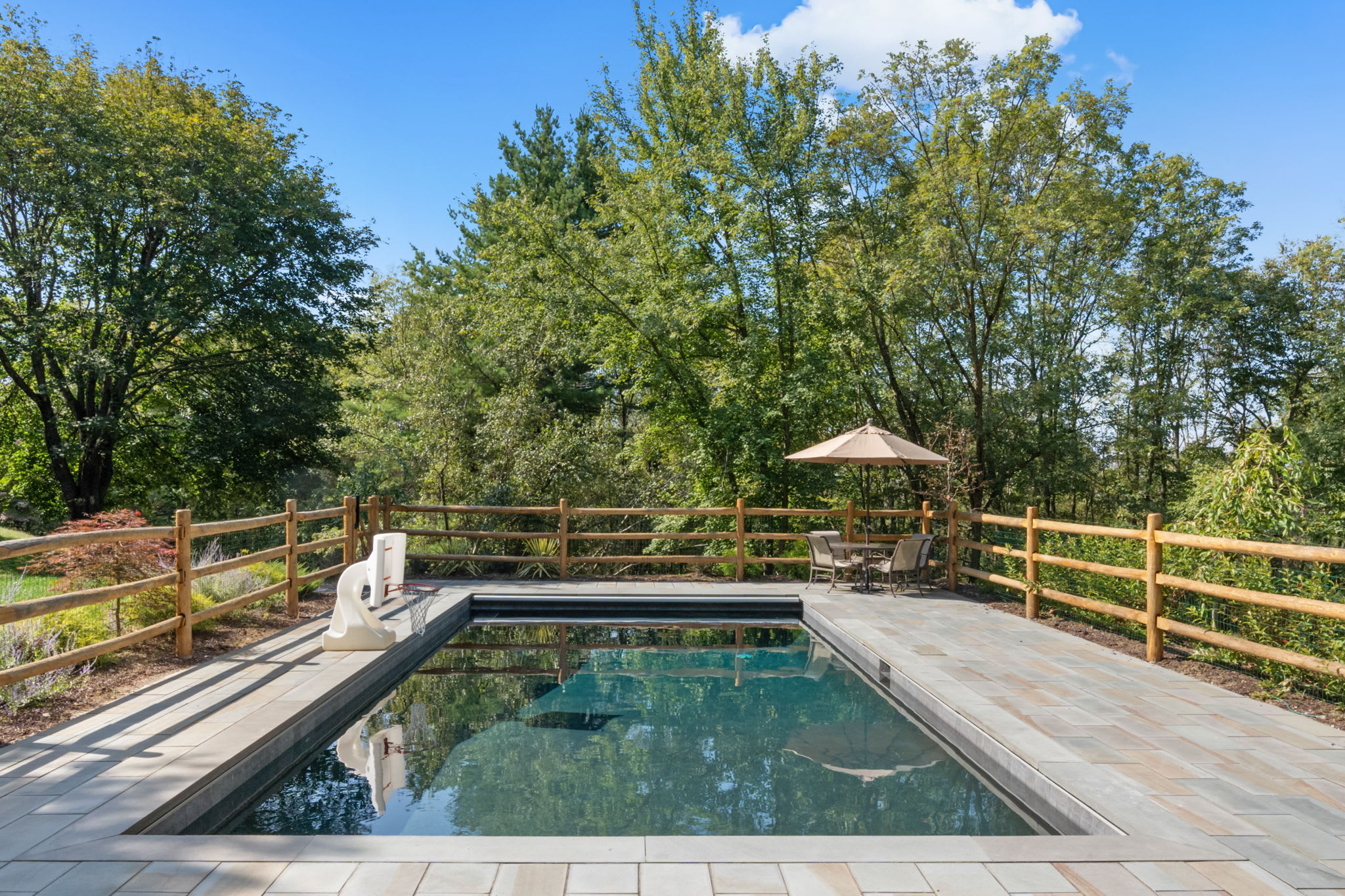 A rectangular inground swimming pool with light gray paver decking is enclosed by a rustic wooden fence, surrounded by lush green trees under a bright blue sky. A small patio area with a table and umbrella is visible at one end of the pool.