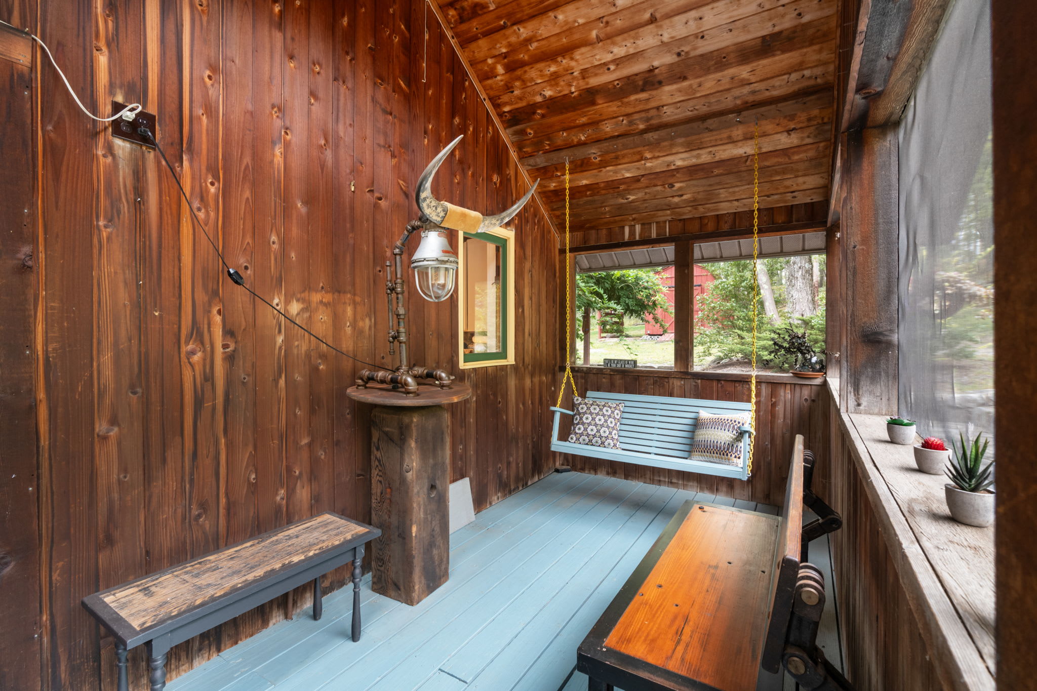 A rustic screened porch with natural wood walls and ceiling, light blue painted floorboards, and a light blue porch swing, adorned with a unique bull horn light fixture and a wooden bench.