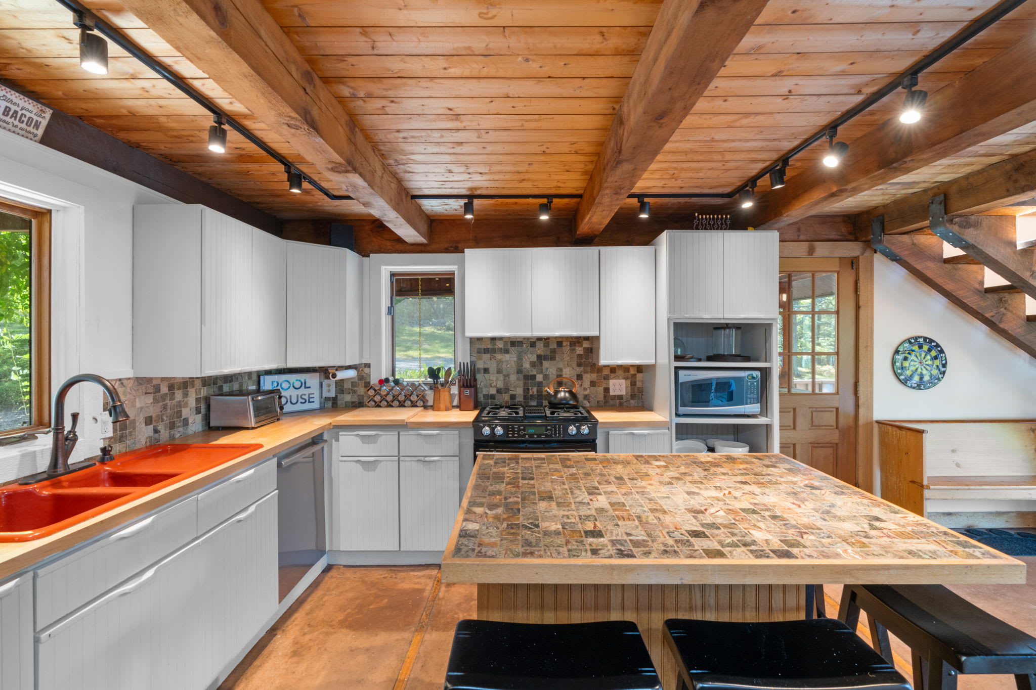 A rustic kitchen with exposed wooden beam ceiling, white shaker cabinets, butcher block countertops, and a central island with a tiled top, equipped with a gas range and stainless steel appliances.