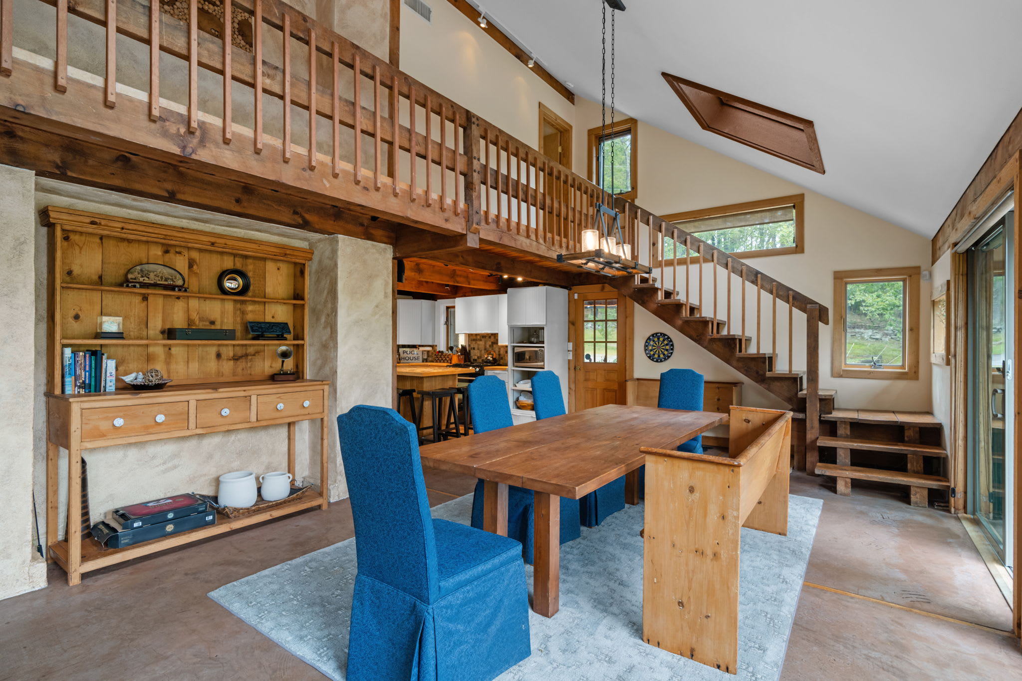 An open-concept dining area features a long wooden table with four blue upholstered chairs and a wooden bench. A wooden railing balcony overlooks the space, with a modern chandelier hanging above the table and a kitchen visible in the background.