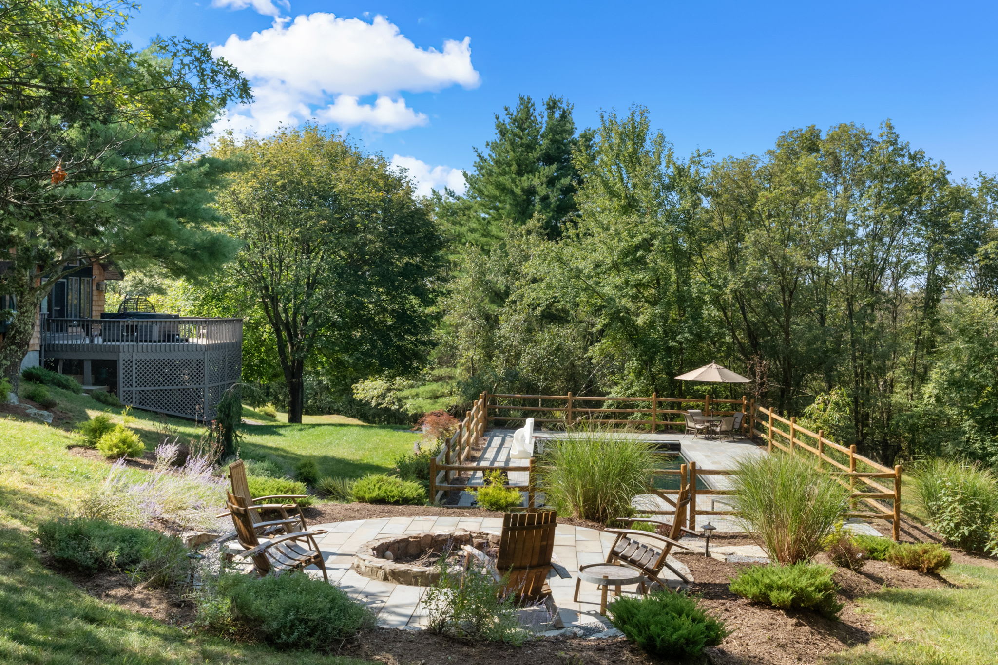 A circular stone fire pit area with multiple wooden Adirondack chairs is nestled on a landscaped terrace, overlooking the fenced swimming pool below. The main house with a deck is visible in the background, surrounded by lush trees.