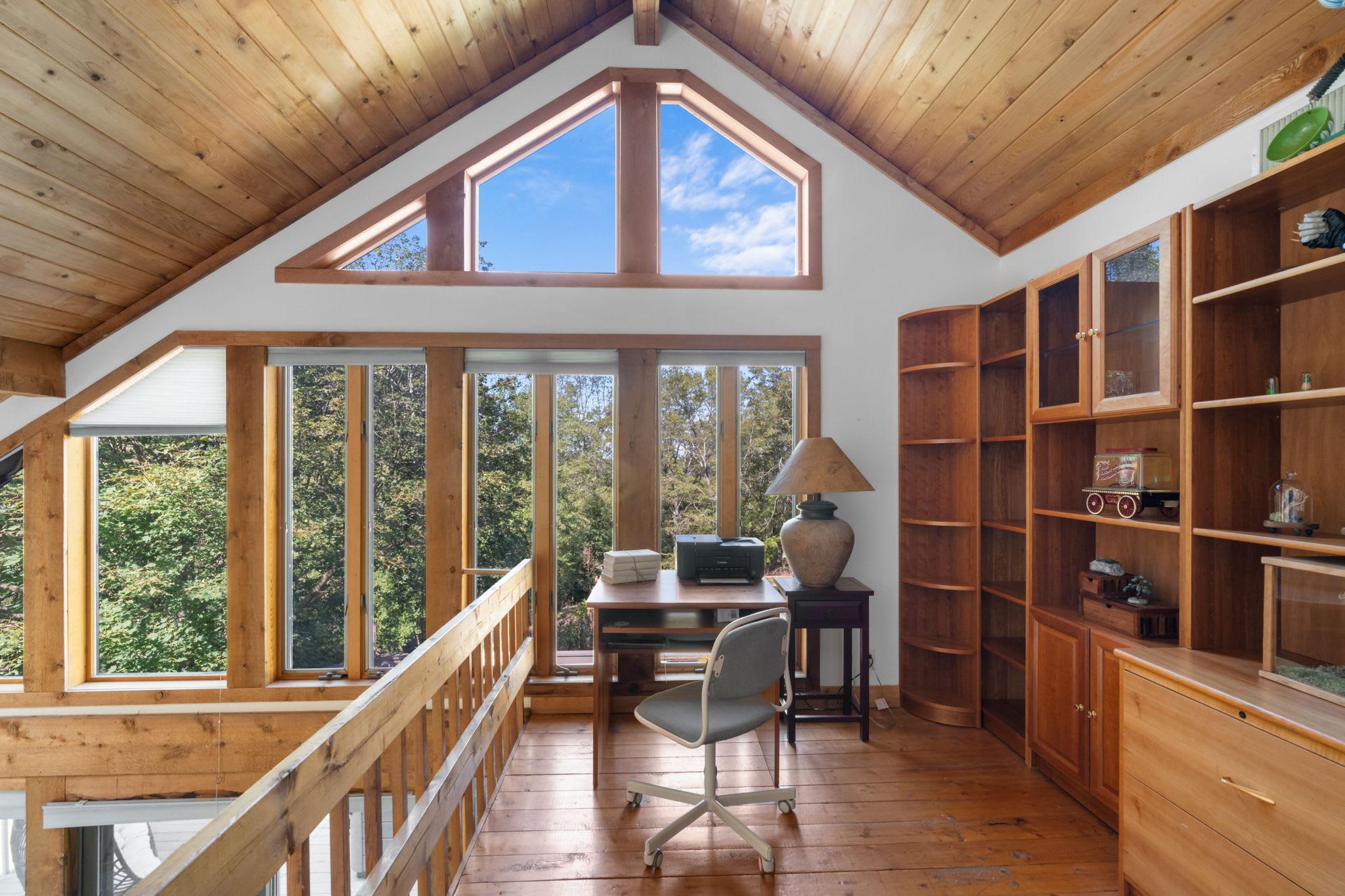 Bright loft office space with a wooden desk and chair, surrounded by built-in wooden bookshelves and large windows offering expansive views of the outdoors, featuring a high vaulted wooden ceiling.