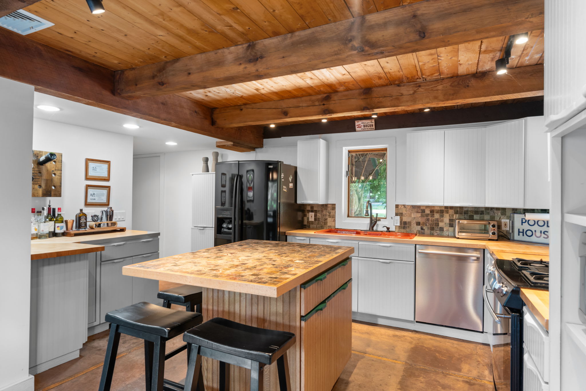 A wide view of the kitchen showcasing white shaker cabinets, butcher block countertops, a black French door refrigerator, and a central island with a tiled top and bar stools, all under a wood beam ceiling.