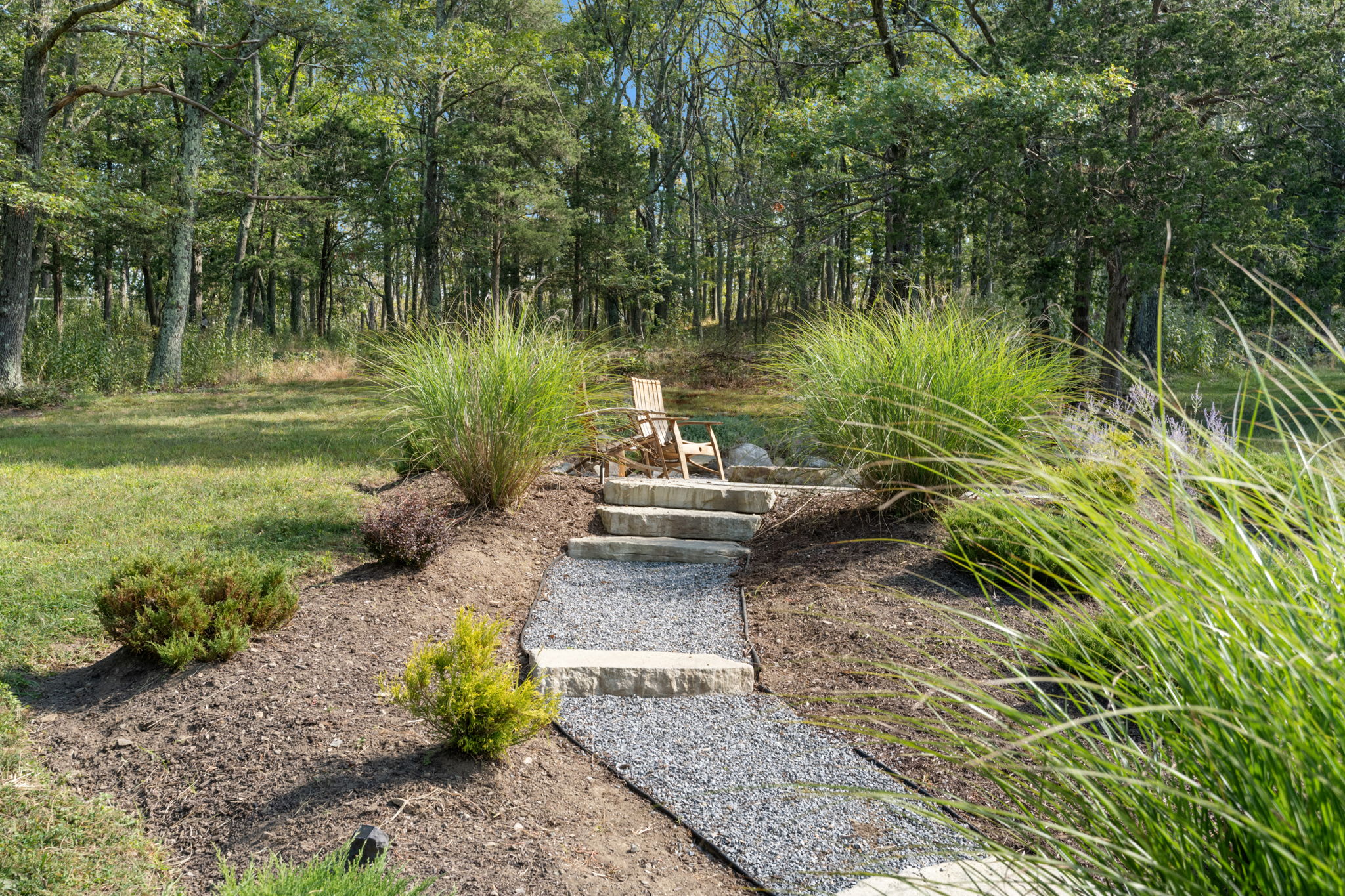 A charming gravel path with large stone steps winds up a landscaped hillside, flanked by ornamental grasses and shrubs. A single wooden Adirondack chair sits at the top of the steps, offering a peaceful spot amidst nature.