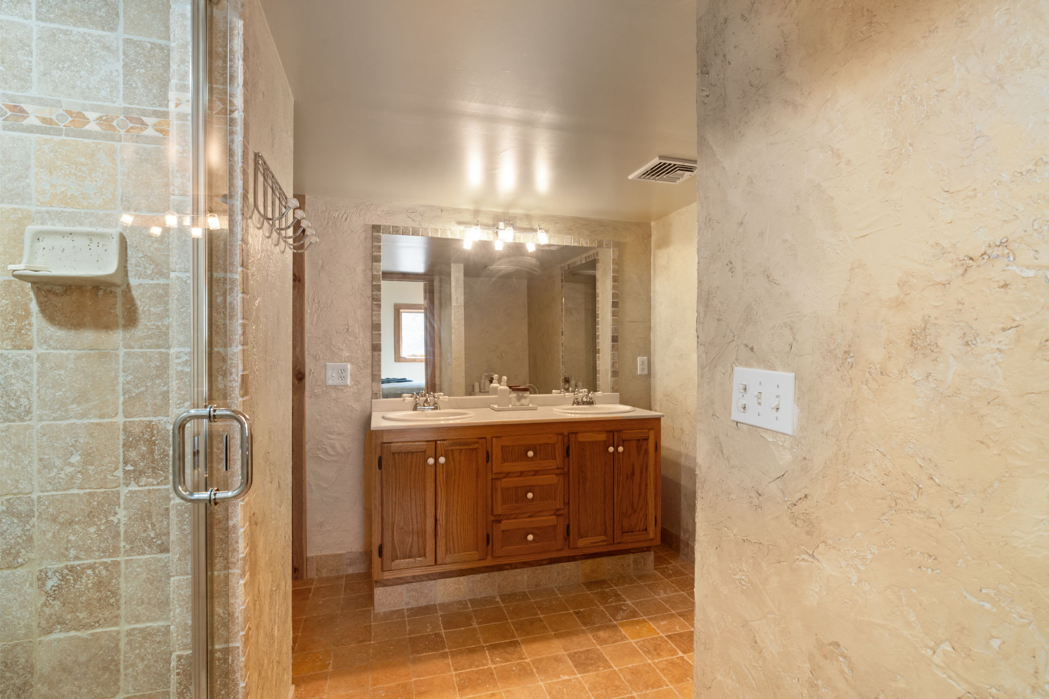 This bathroom features a double vanity with a wooden cabinet and dual sinks, illuminated by a large mirror and bright vanity lighting. The floor is covered in warm, earthy-toned tiles, complementing the textured walls.