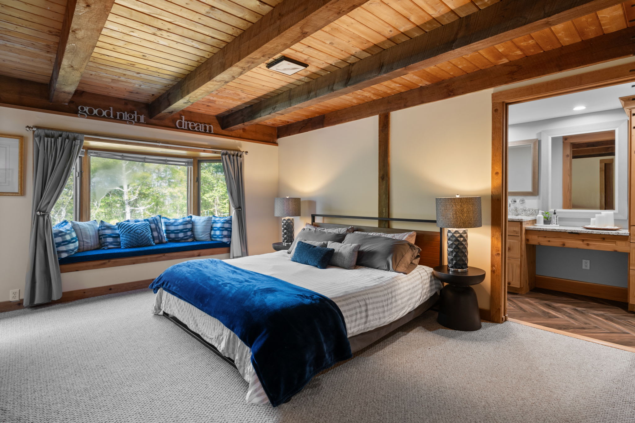 A cozy bedroom features a queen-sized bed with blue and gray bedding, flanked by two nightstands with lamps. A large bay window with a built-in window seat and blue pillows offers views of trees, complemented by a vaulted wooden beam ceiling.
