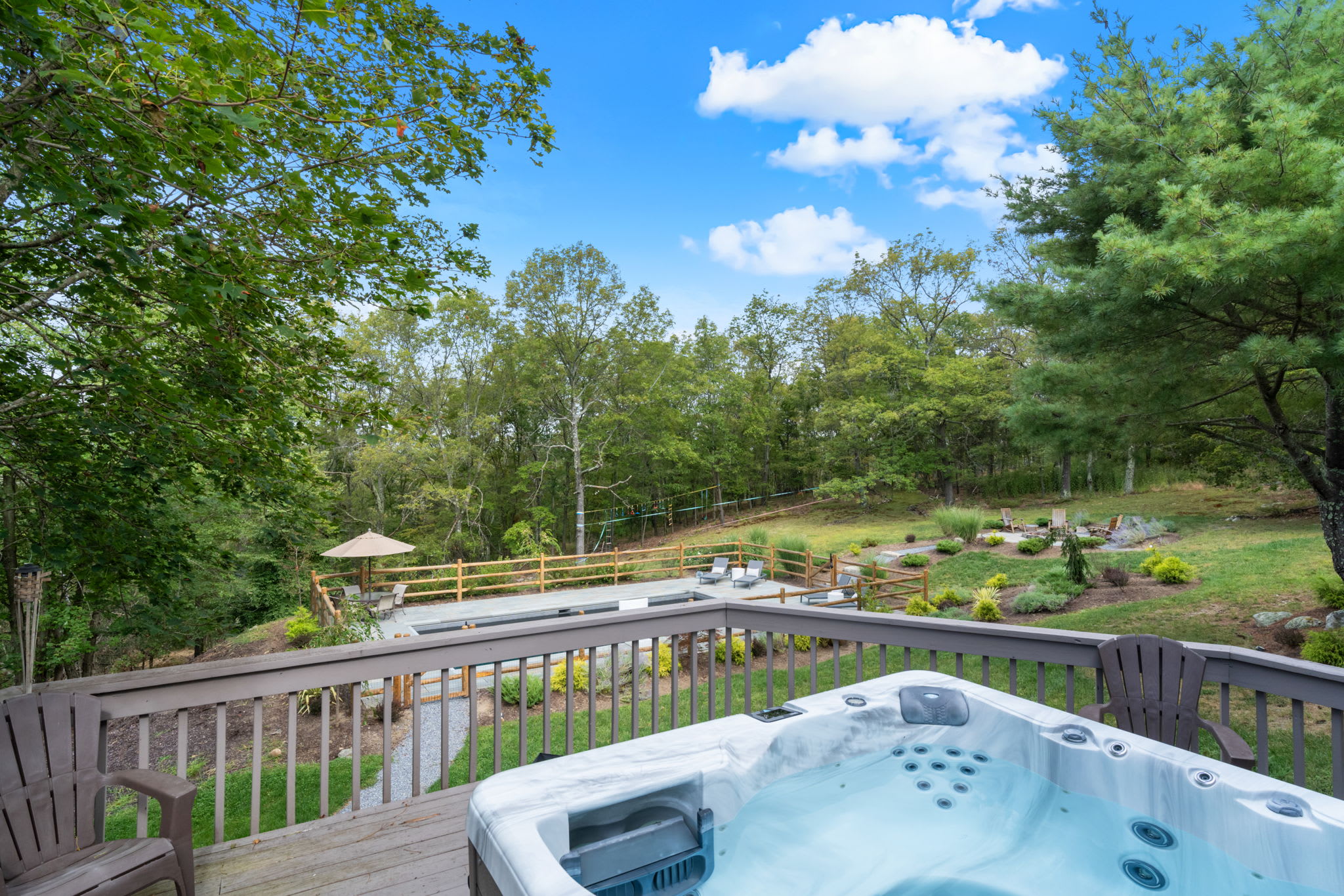 An inviting outdoor hot tub is situated on a wooden deck, offering a relaxing spot with views of a fenced swimming pool area and a sprawling green yard surrounded by mature trees under a bright blue sky.