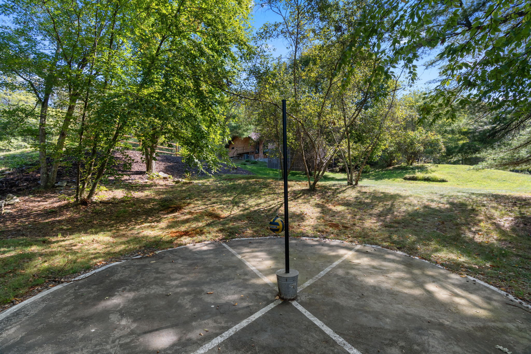 A concrete four-square court with a central pole and a tetherball, set in a sun-dappled grassy yard surrounded by lush green trees. The court shows signs of wear with cracks and moss, indicating a well-used outdoor play area.
