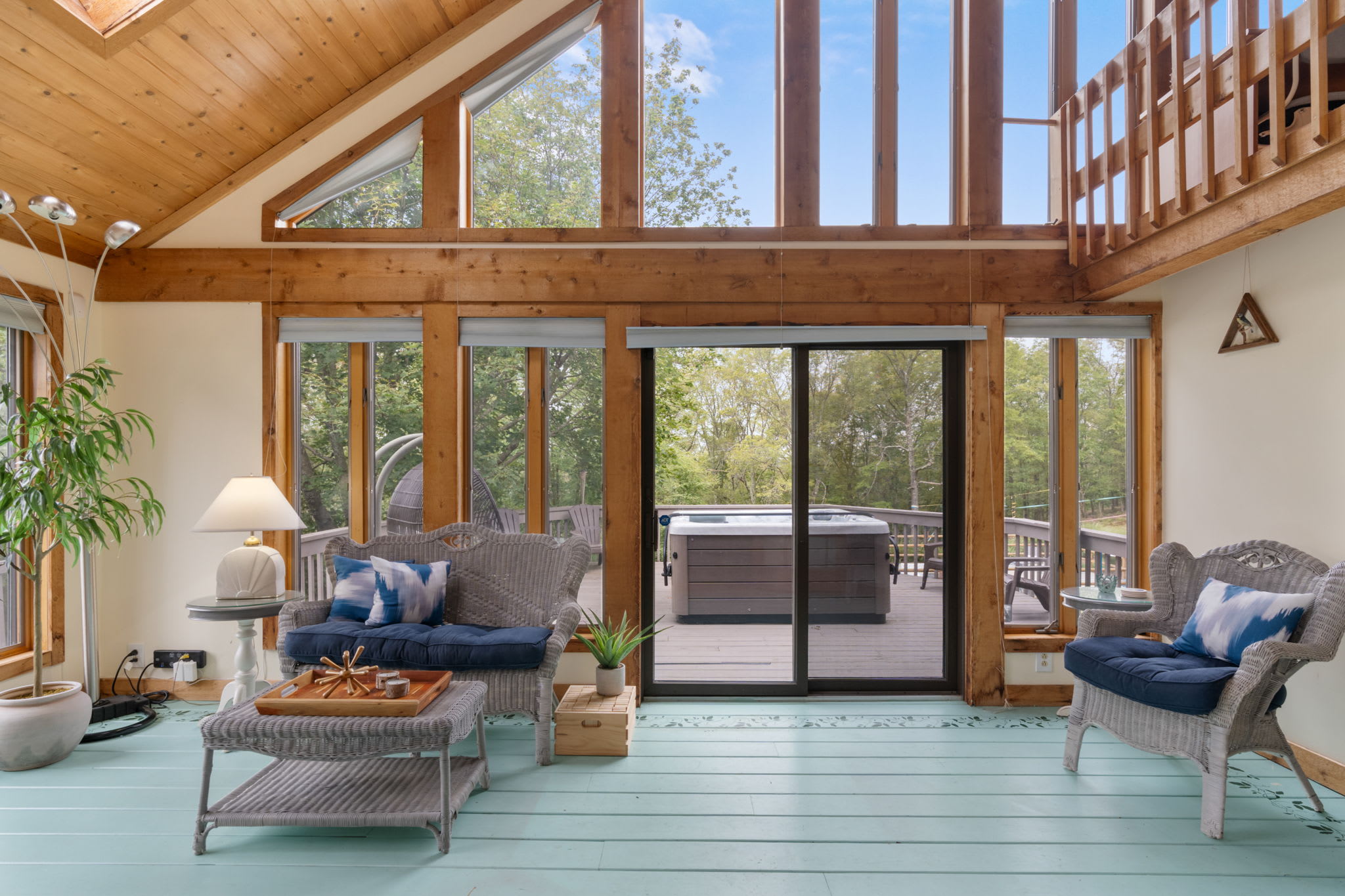 Bright sunroom with light blue painted floorboards, wicker furniture with blue cushions, and large windows offering extensive views of the surrounding trees and access to the hot tub deck.