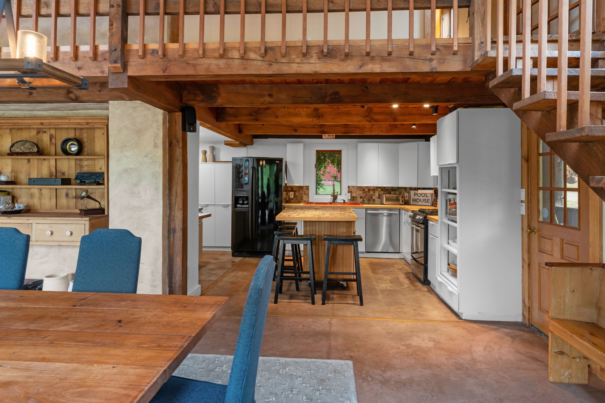 An open-concept kitchen and dining area featuring exposed wooden beams, white cabinetry, a central island with bar seating, and a large wooden dining table with blue chairs.