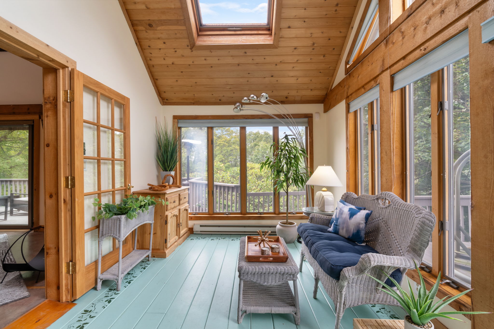 A bright sunroom with a vaulted wood ceiling and skylight, featuring light blue painted floorboards with a decorative stencil, wicker furniture, and large windows offering expansive outdoor views.