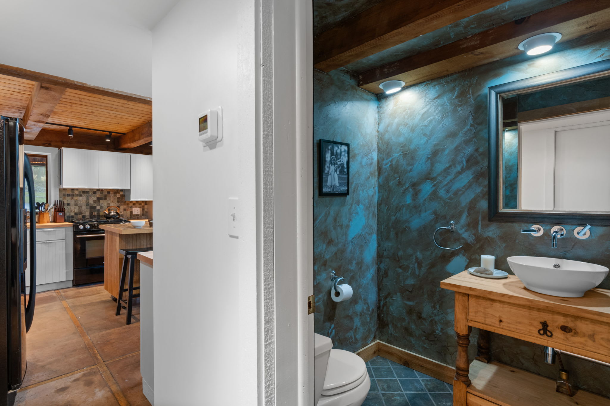 A compact powder room featuring a unique blue textured wall, a rustic wooden vanity with a vessel sink, and a toilet, illuminated by recessed lighting and an exposed wood beam.