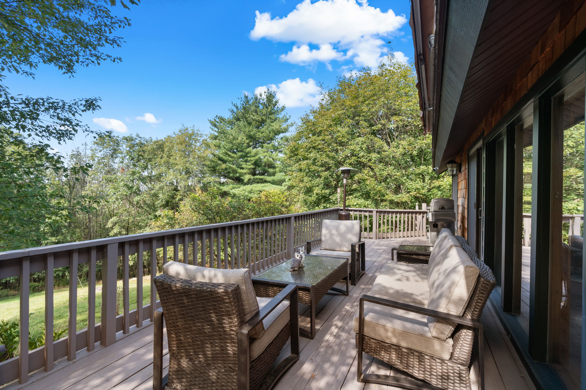 A spacious outdoor deck is furnished with comfortable wicker seating, including a sofa and armchairs, arranged around a glass-top coffee table. A patio heater stands nearby, and the deck offers scenic views of lush green trees under a blue sky.