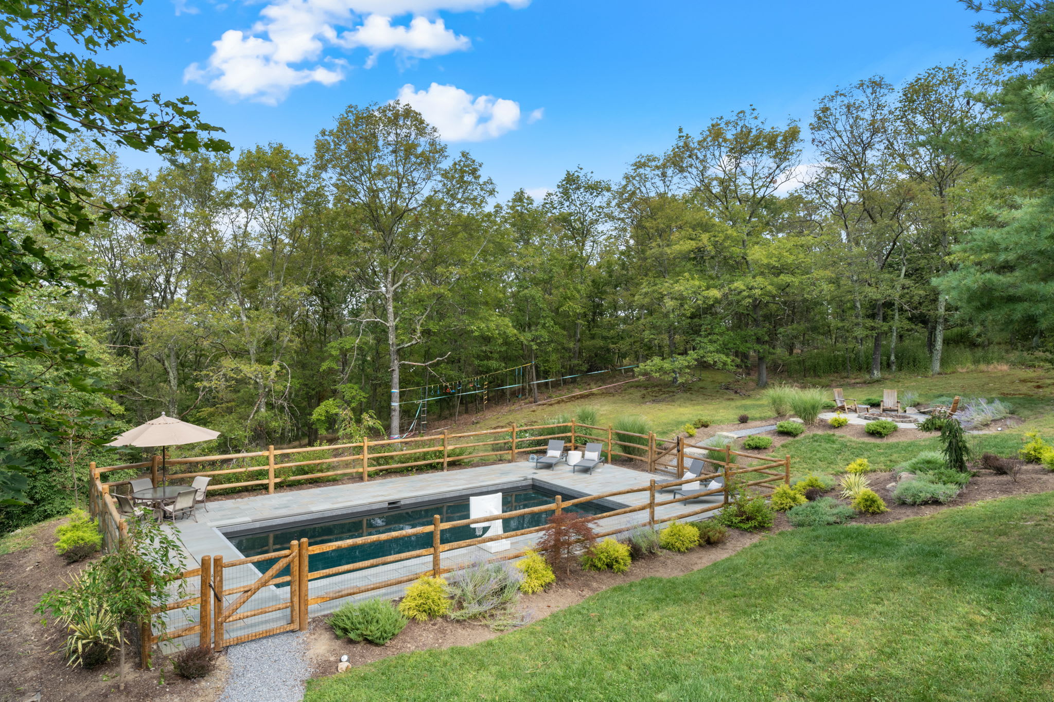 An elevated view shows a rectangular inground pool with gray paver decking, enclosed by a rustic wooden fence, set into a sloped grassy yard. Beyond the pool, a fire pit area and dense forest are visible under a clear blue sky.