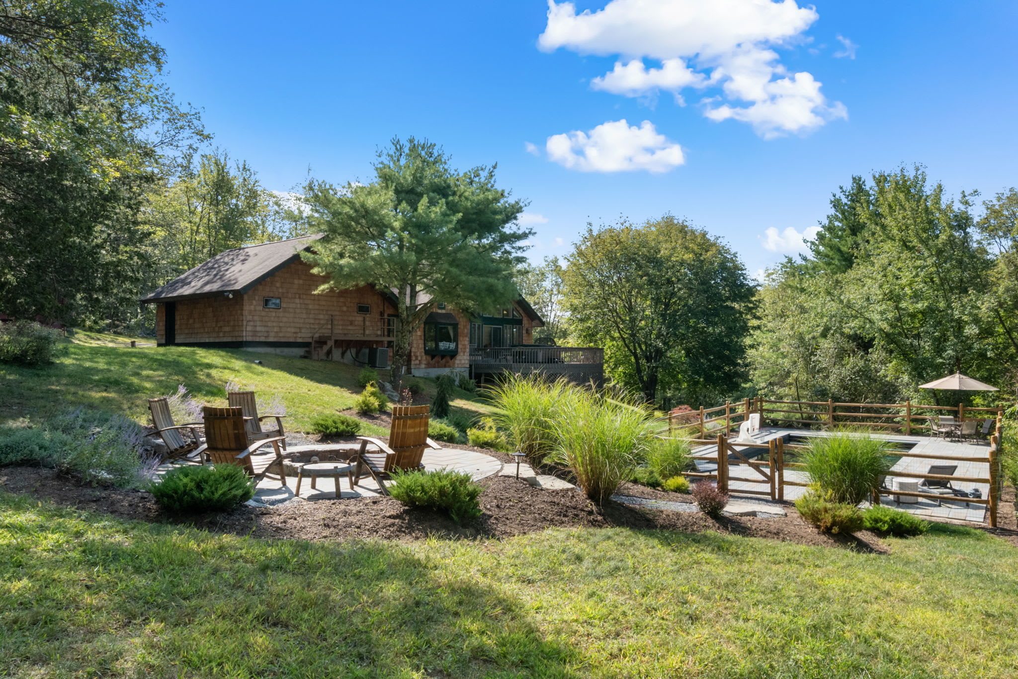 The back exterior of a rustic cabin-style house with cedar siding and a large deck is visible from a grassy hillside, overlooking a fire pit area and a fenced swimming pool. The property is surrounded by dense, mature trees under a bright blue sky.