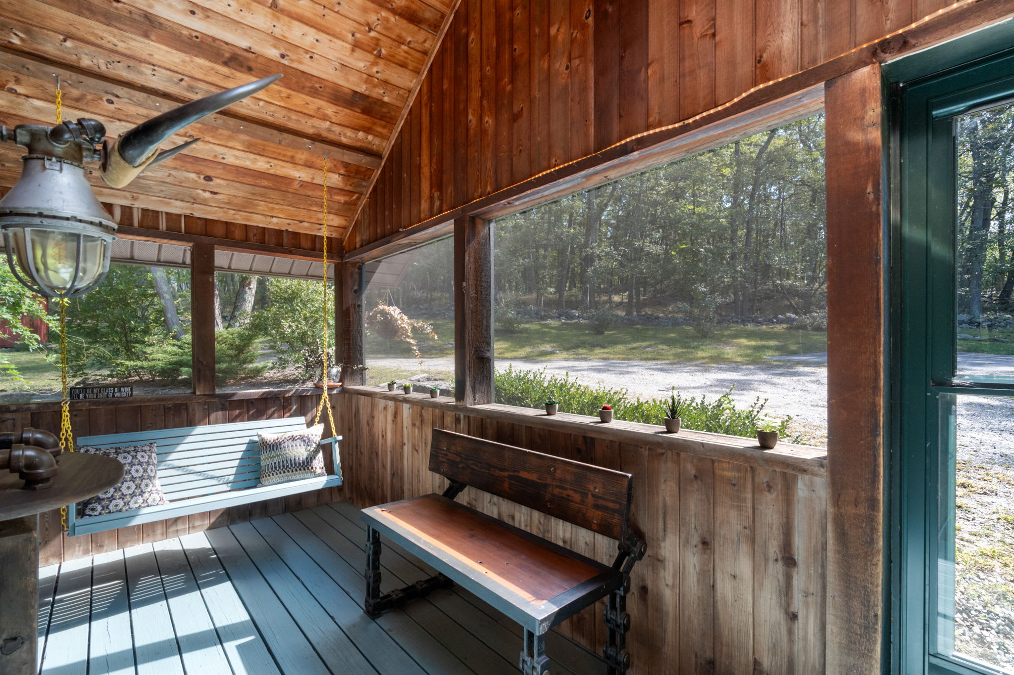 A cozy screened porch with light blue painted floorboards and a matching swing, featuring natural wood walls and ceiling, offering views of the gravel driveway and surrounding trees.