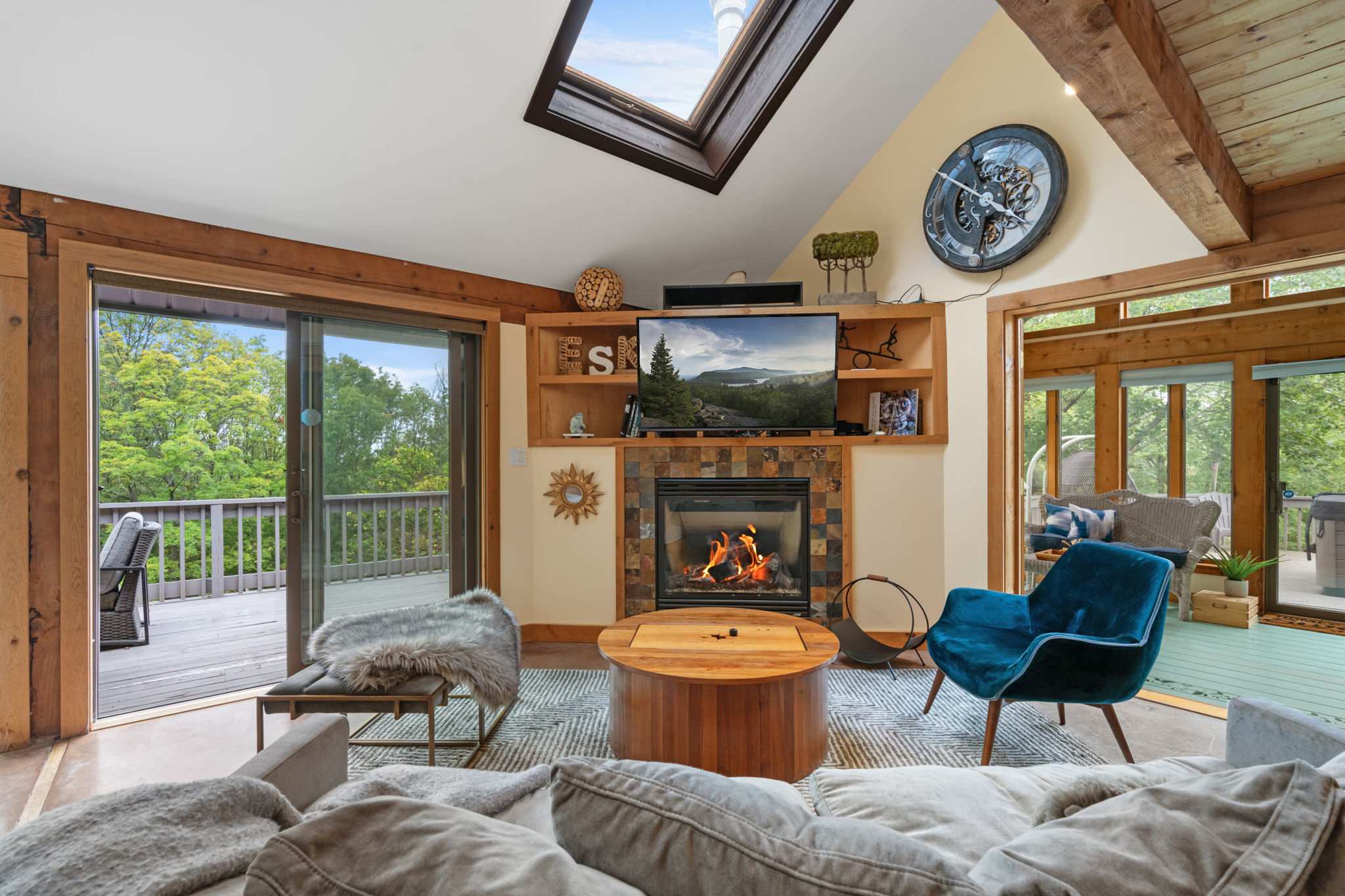 Cozy living room with a stone-tiled fireplace, wall-mounted TV, and a round wooden coffee table, featuring a skylight and sliding glass doors leading to an outdoor deck.