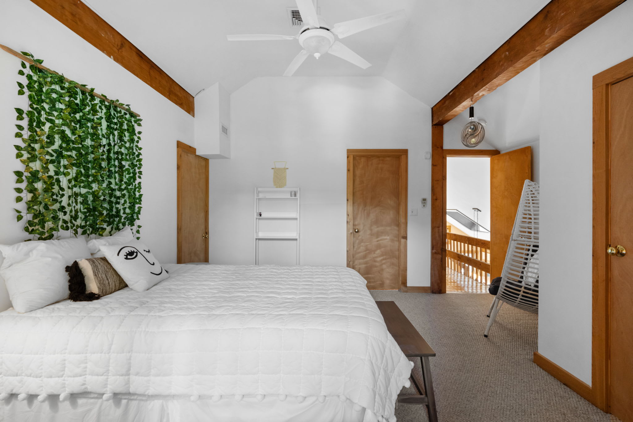 This view of bedroom-3 highlights the queen bed with white bedding, a unique white hanging chair, and a white shelving unit. The room's vaulted ceiling with a fan and exposed wooden beams adds to its rustic charm, with a doorway leading to an upper-level hallway.