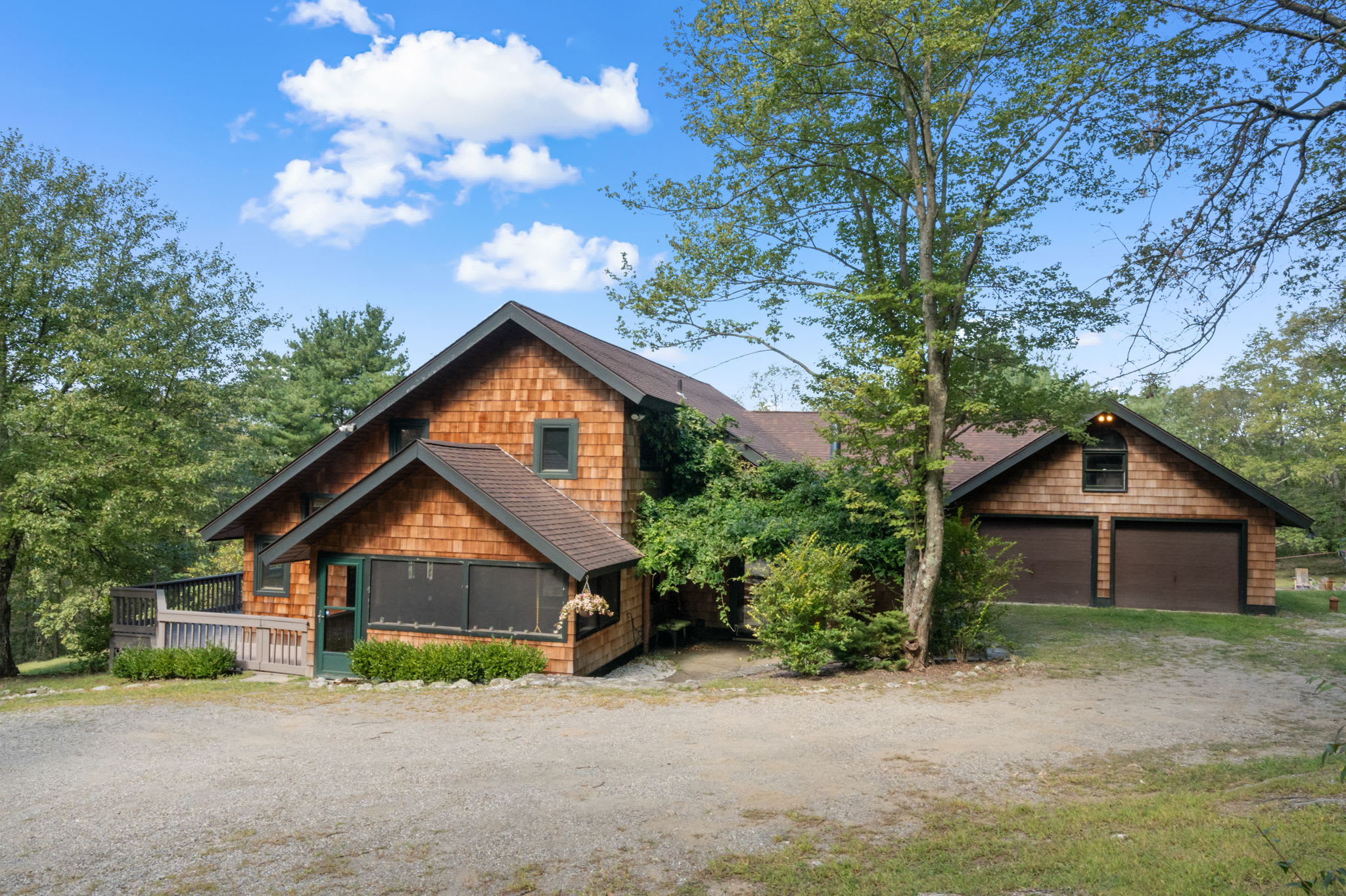 A rustic, multi-level cabin with cedar shake siding and dark trim, featuring a screened porch and an attached two-car garage, surrounded by lush green trees under a blue sky.