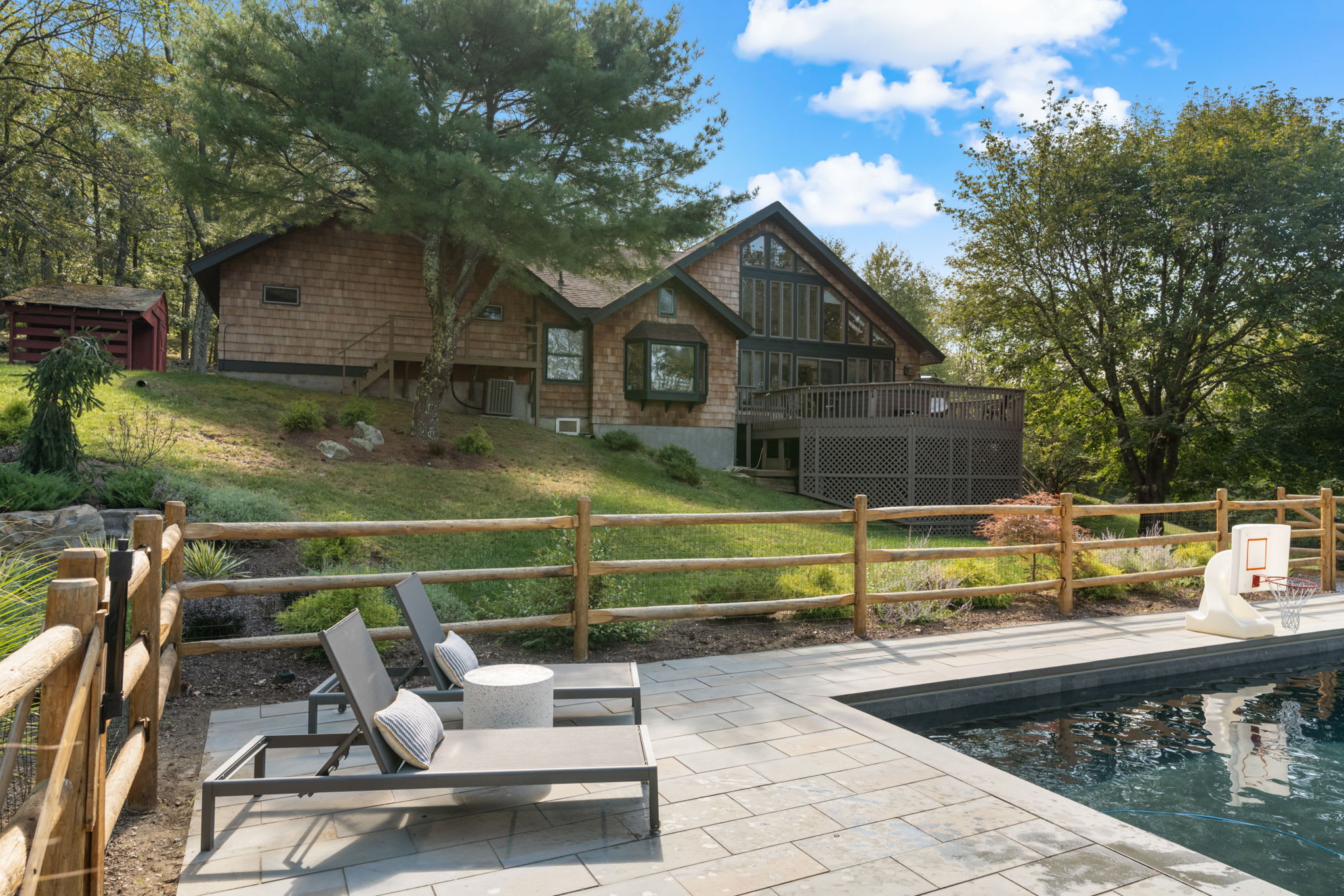 A rectangular inground swimming pool with light gray paver decking is enclosed by a rustic wooden fence, surrounded by lush green trees under a bright blue sky. A small patio area with a table and umbrella is visible at one end of the pool.