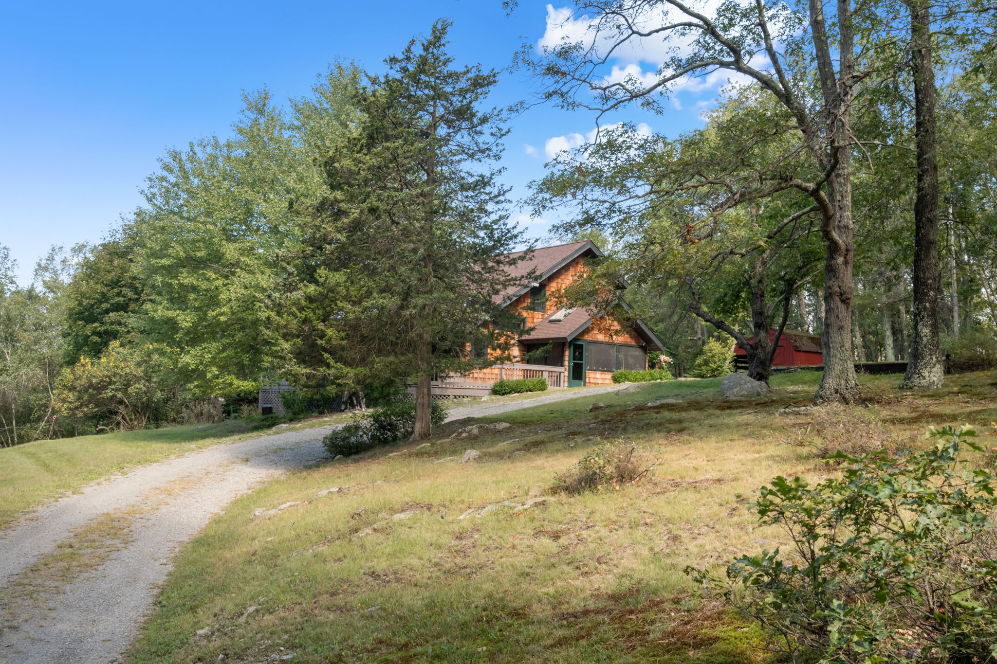 A rustic, multi-story cabin with warm wood siding and a dark roof, nestled among mature trees on a sloped, grassy landscape. A gravel driveway curves towards the house, with a small red outbuilding visible in the distance.