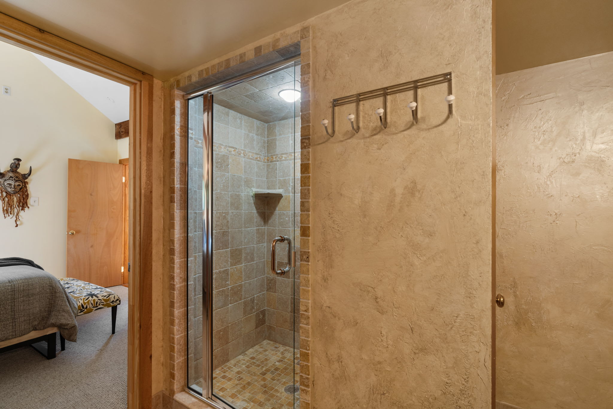 A spacious walk-in shower with a glass door and built-in shelf is clad in light-colored stone-look tiles. The adjacent wall features a textured finish and a metal coat rack with white knobs.