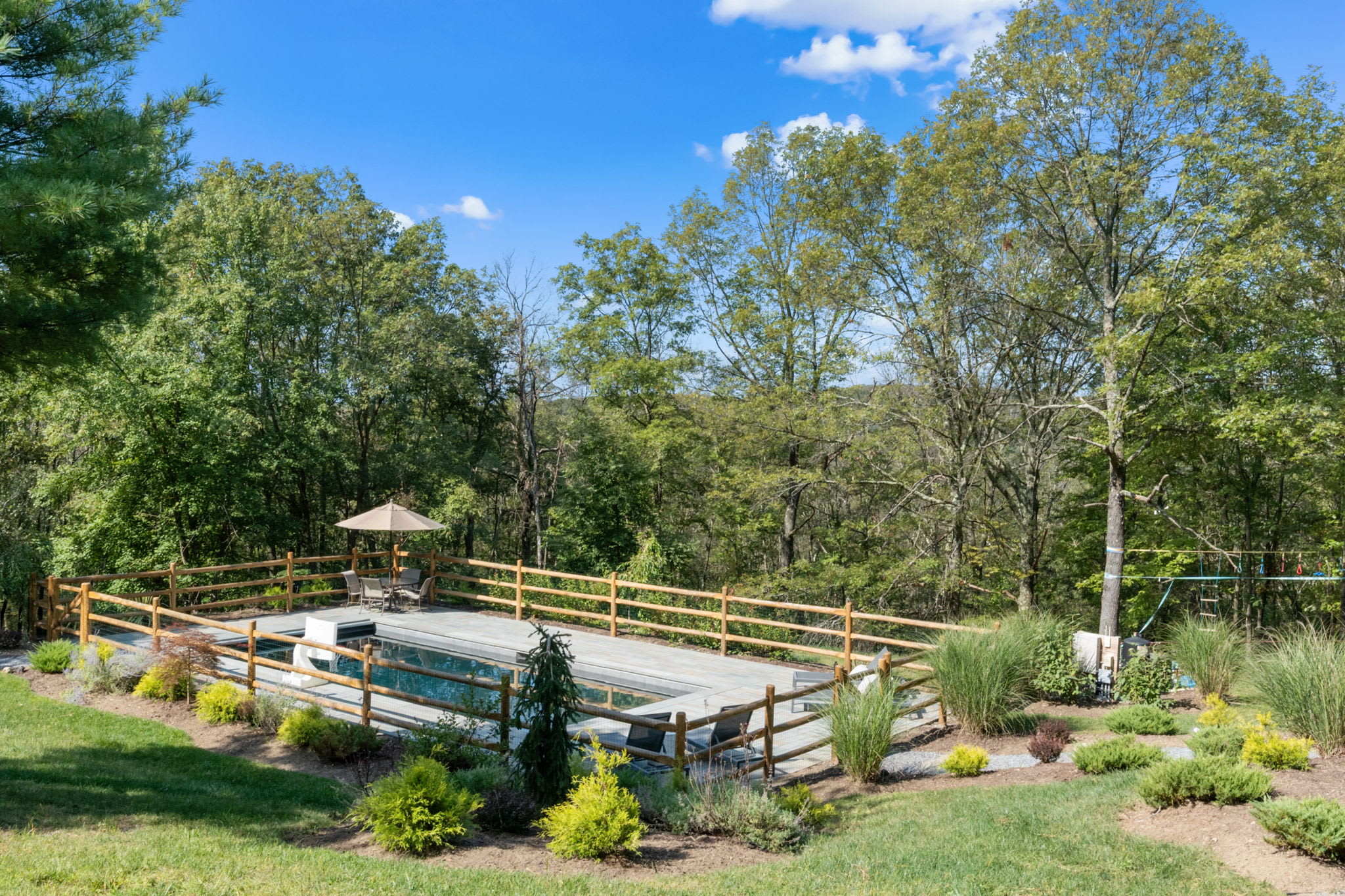 An elevated view captures the rectangular inground pool with gray paver decking, enclosed by a rustic wooden fence, nestled in a landscaped, sloped yard. Beyond the pool, a vast expanse of green forest stretches under a clear blue sky.